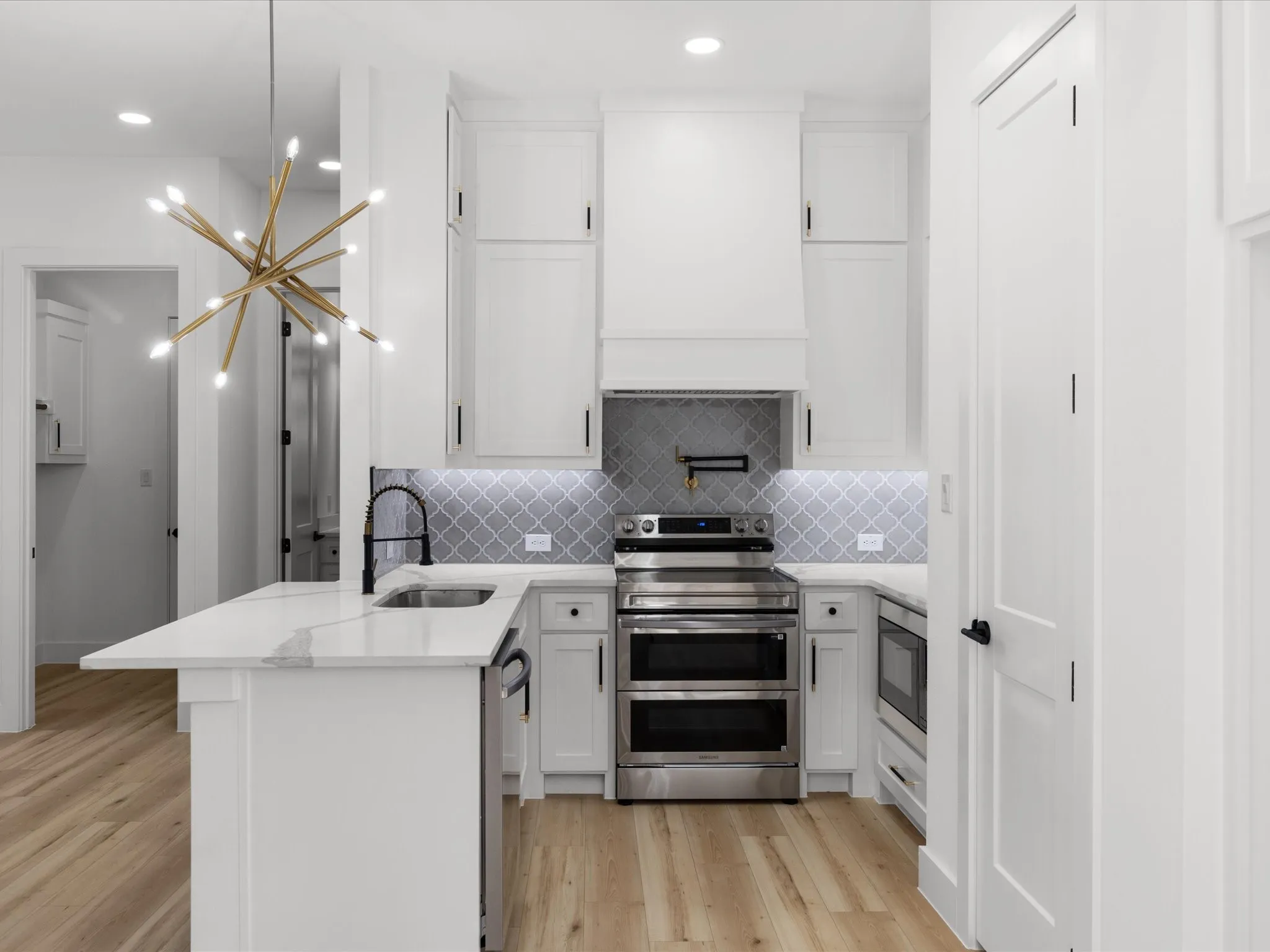 Modern kitchen featuring white cabinetry, light-toned wood-finish flooring, stainless steel appliances, a white countertop island with an undermount sink, and a gray arabesque tile backsplash