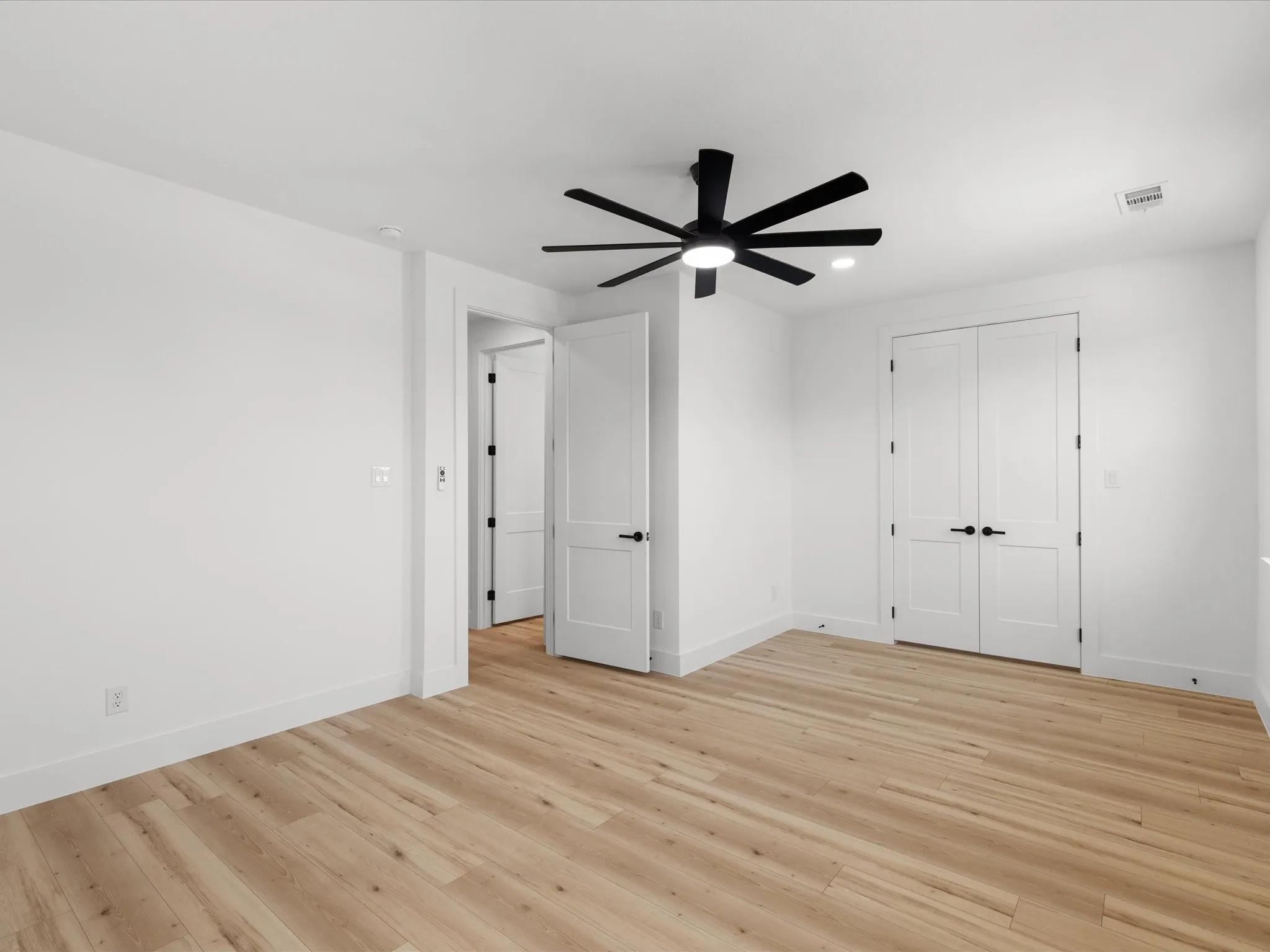 Bright interior space featuring light wood-finish flooring, a modern black ceiling fan with integrated lighting, recessed ceiling light fixtures, white baseboards, and white double doors with black hardware