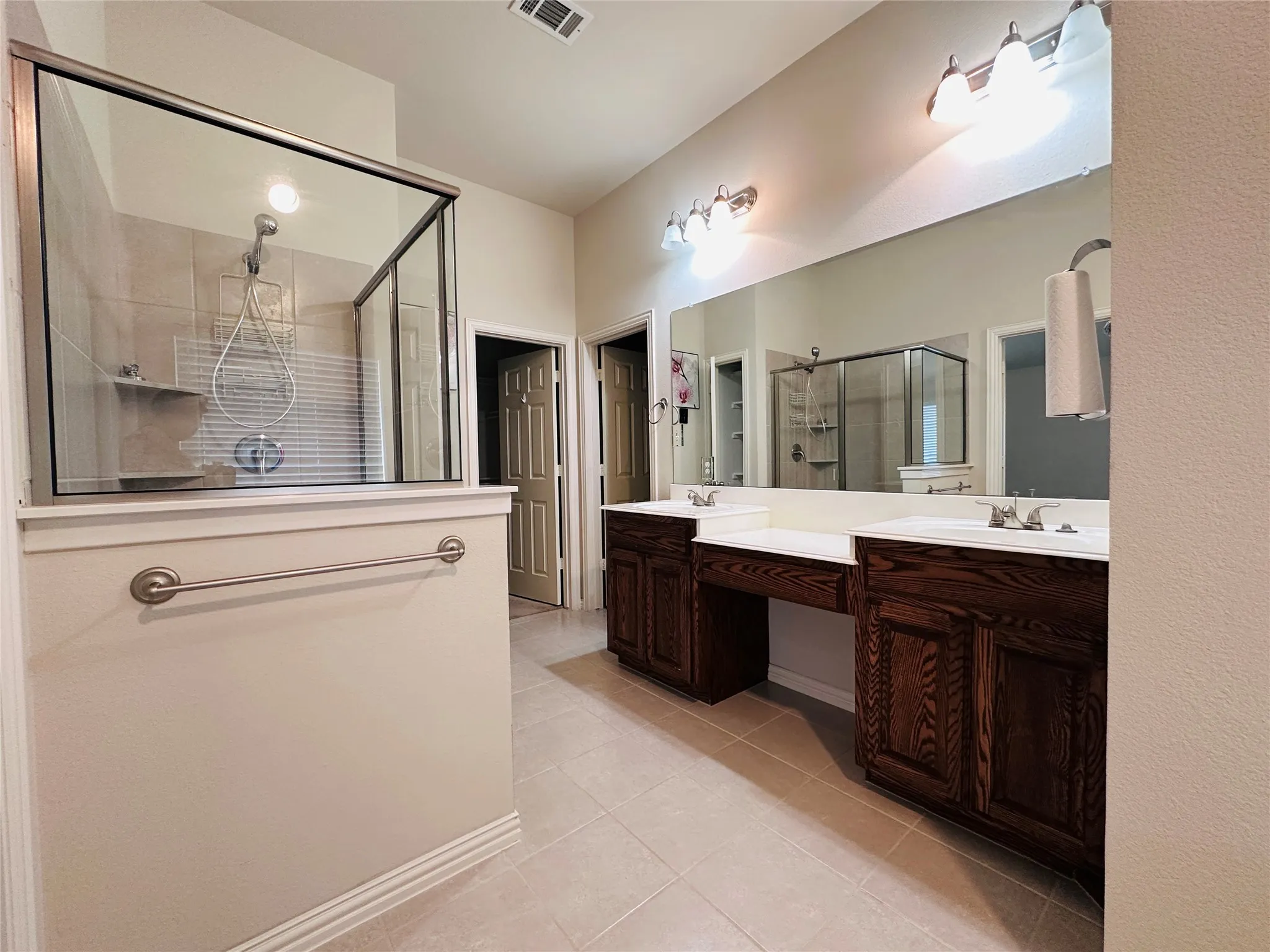 Spacious bathroom featuring a glass-enclosed shower, dual dark wood-finish vanity cabinets, a large wall-mounted mirror, and tile flooring