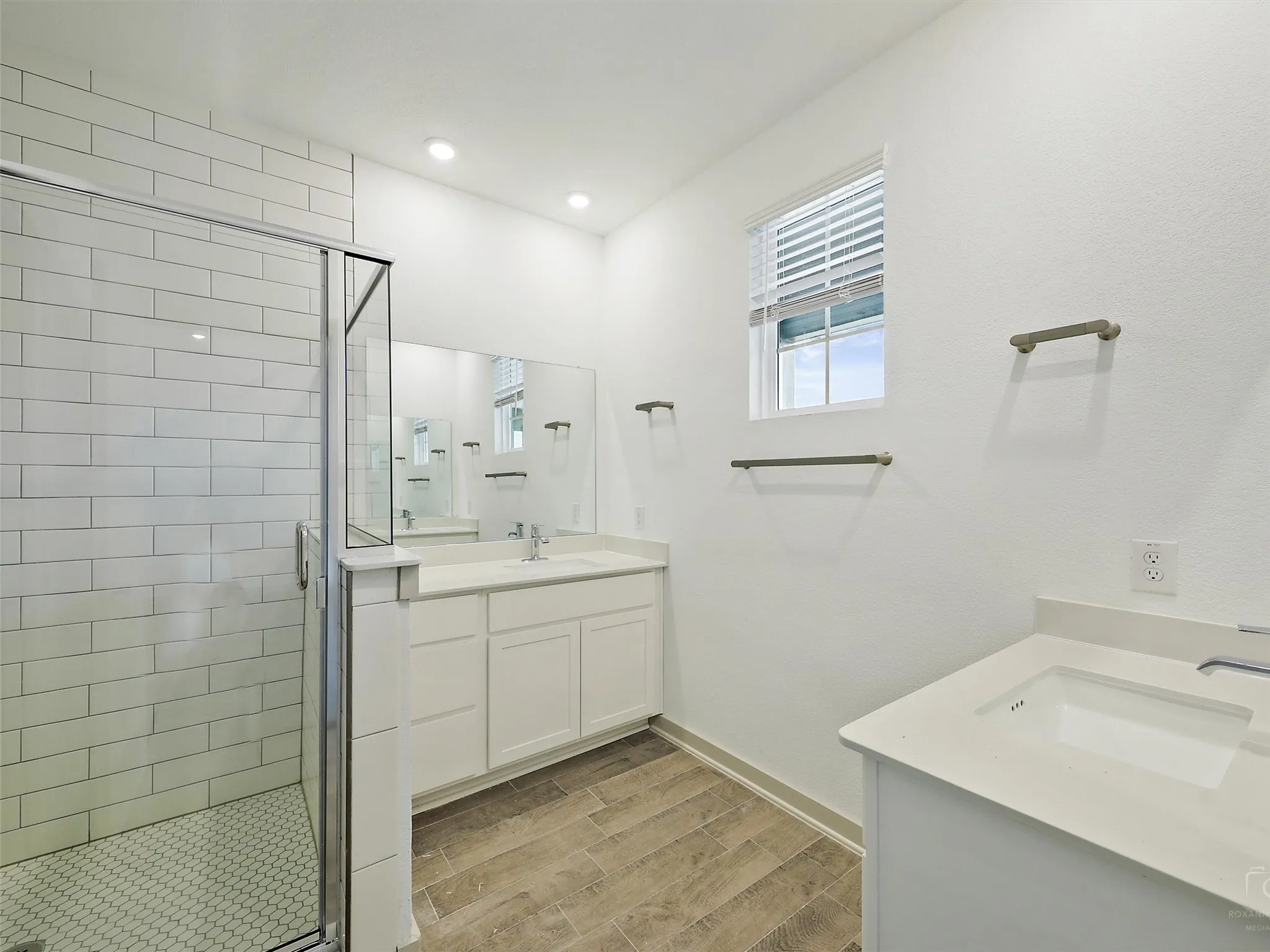 Bathroom featuring a glass-enclosed shower with subway tile surround and hexagonal tile floor, dual vanity sinks with white countertops and cabinetry, wood-finish flooring, and a window with blinds