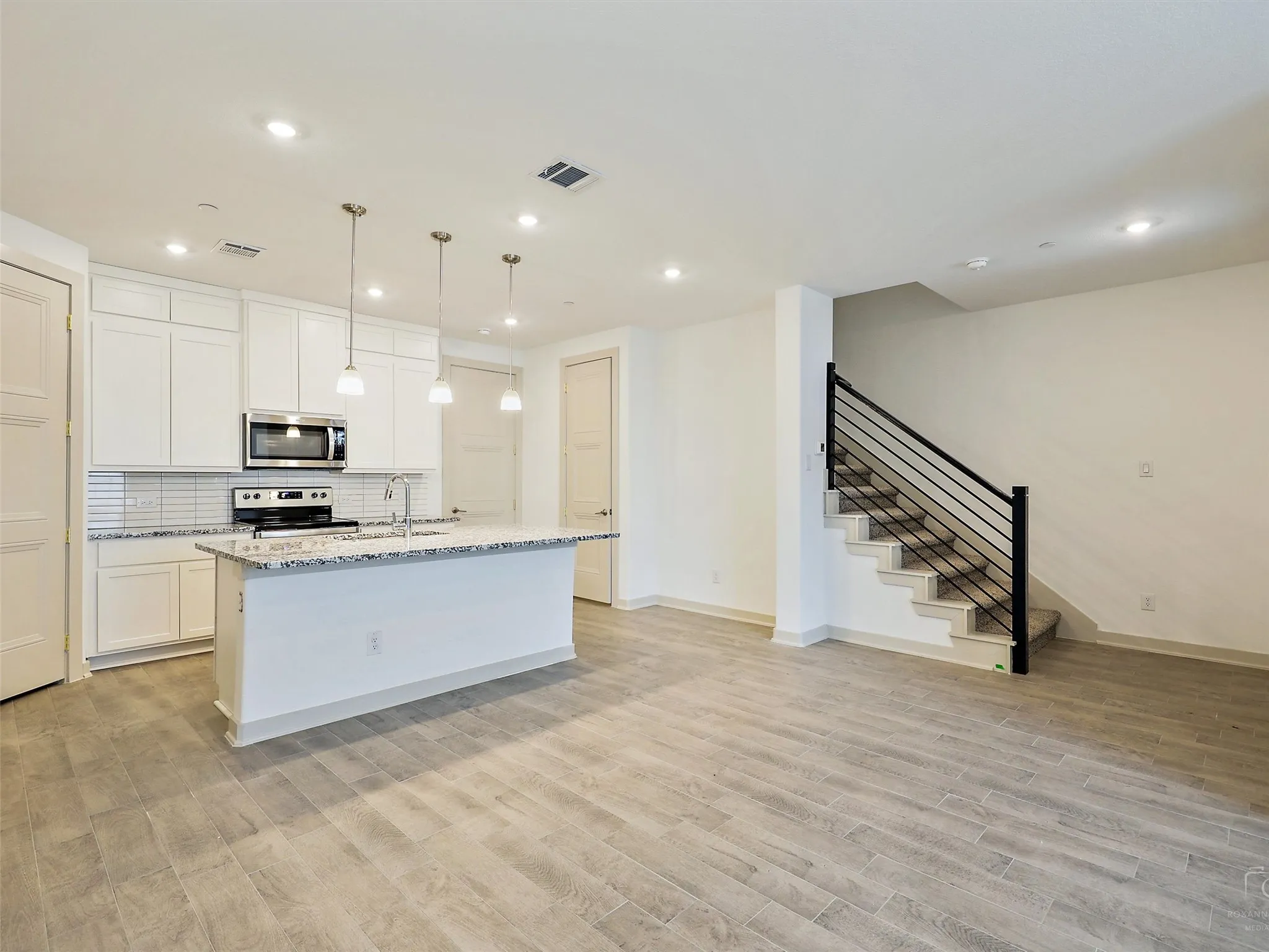 Open-concept kitchen and living area featuring wood-finish flooring, white cabinetry, a kitchen island with seating, stainless steel appliances, and a modern staircase with a black metal railing