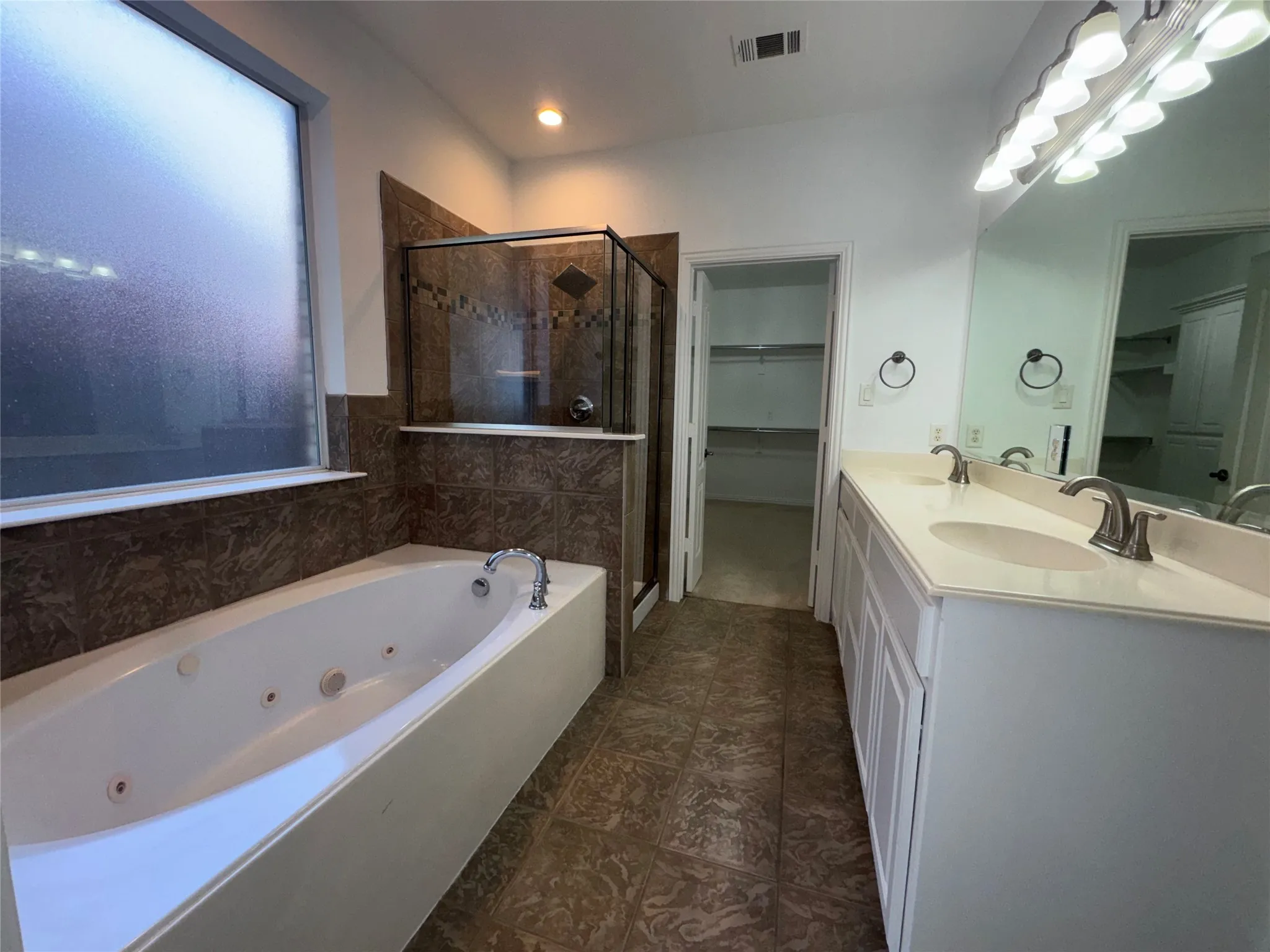 Spa-inspired master bathroom featuring a soaking tub with glass enclosure and tile surround, dual vanities with white cabinetry and undermount sinks, large framed mirror with integrated lighting, and dark tile flooring throughout
