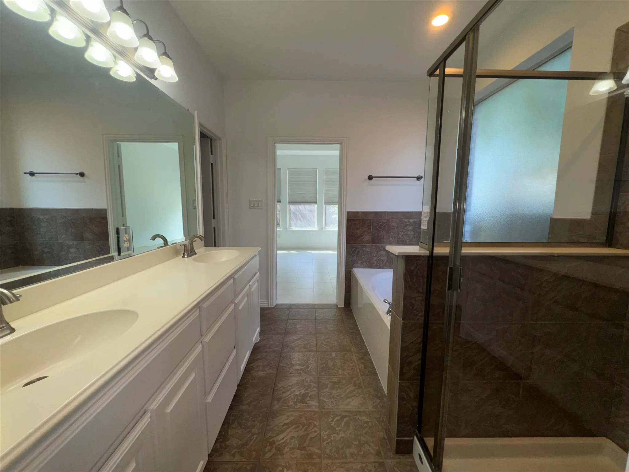 Bathroom featuring a dual vanity with white cabinetry and integrated sinks, a large wall-mounted mirror, a glass-enclosed shower, a soaking tub, and floor-to-ceiling tile accents