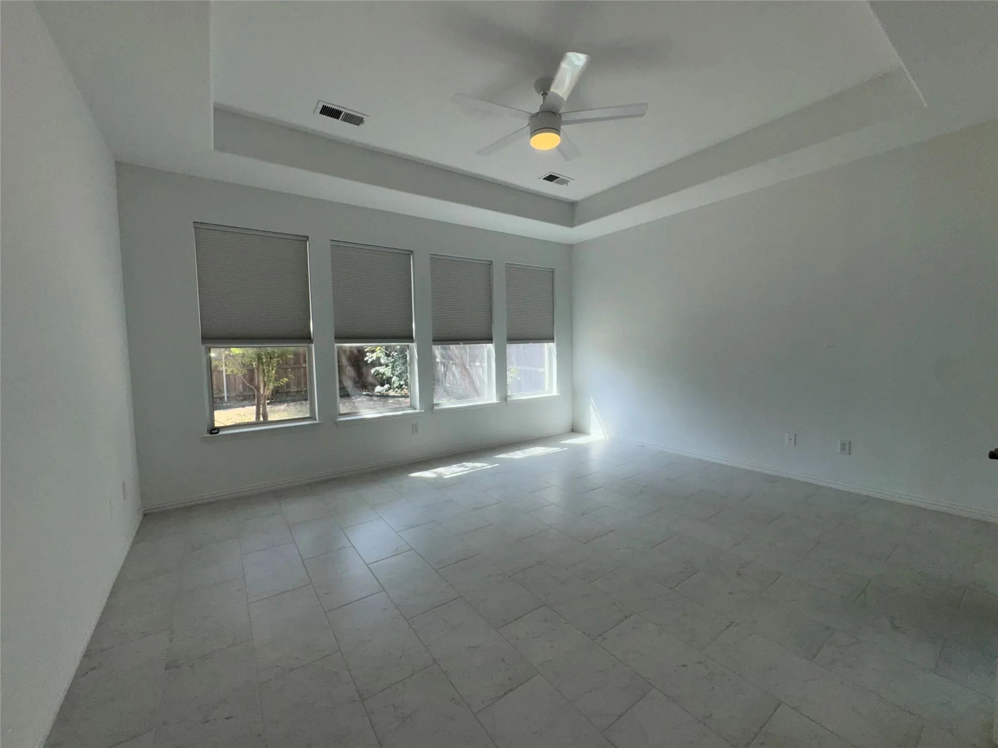 Spacious interior featuring a tray ceiling with a modern ceiling fan, large rectangular tile flooring, and a wall of three windows with roller shades