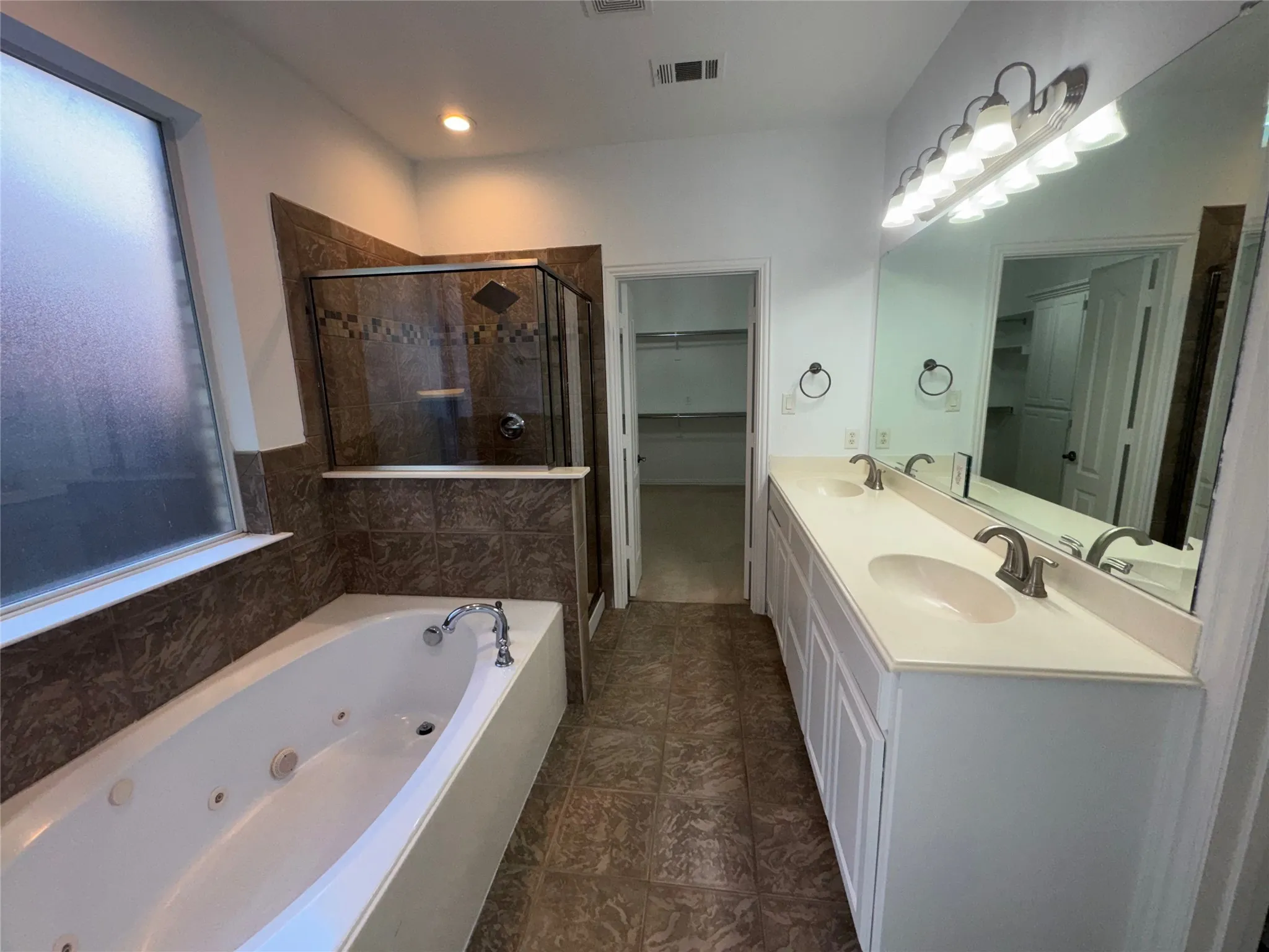 Spa-inspired master bathroom featuring a luxurious whirlpool soaking tub with glass enclosure, dual-sink vanity with white cabinetry and brushed nickel fixtures, large framed mirror with integrated lighting, and wood-finish flooring throughout