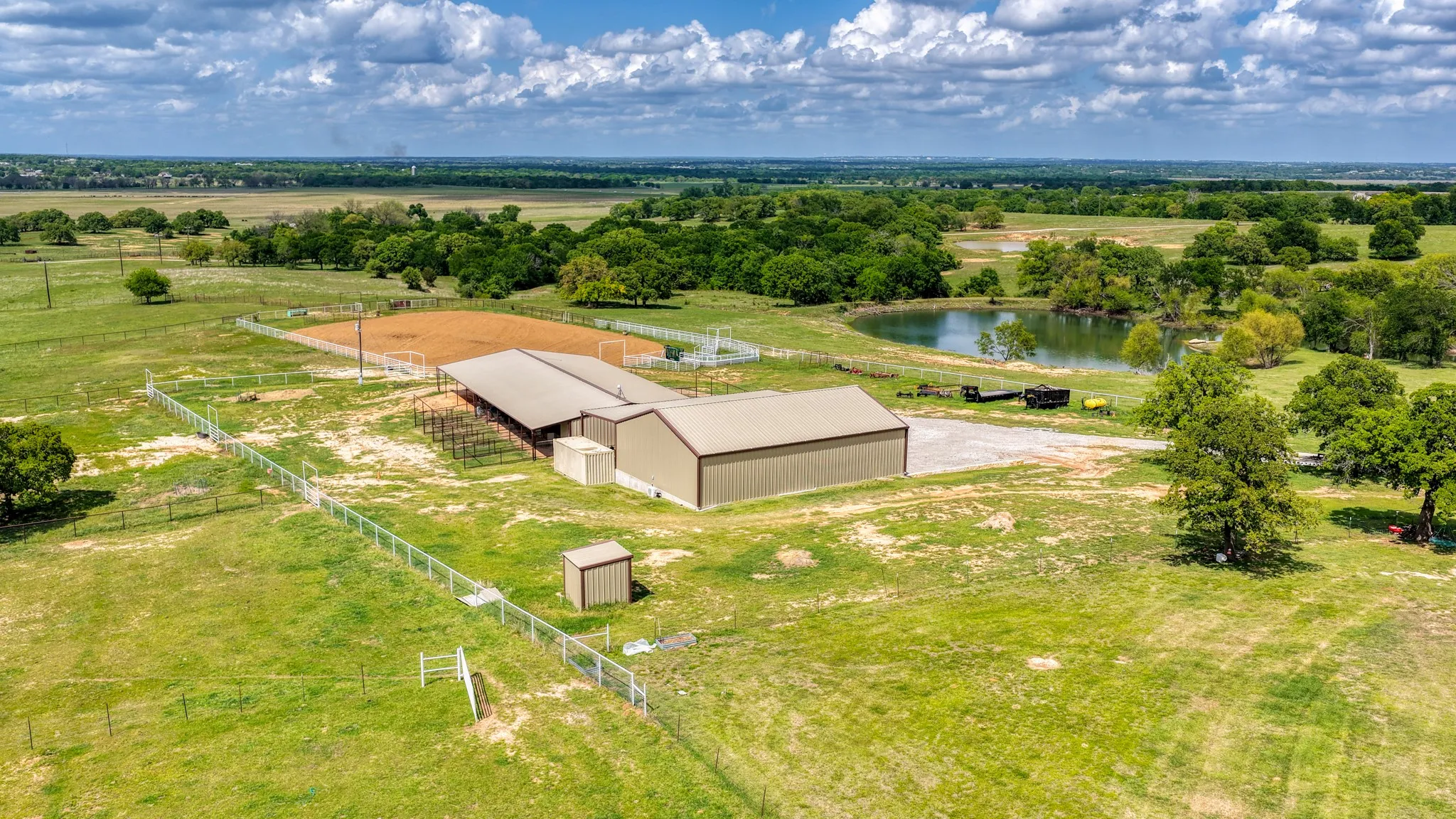 Expansive rural property featuring multiple metal buildings, a fenced riding arena, a separate fenced pasture, and a water feature