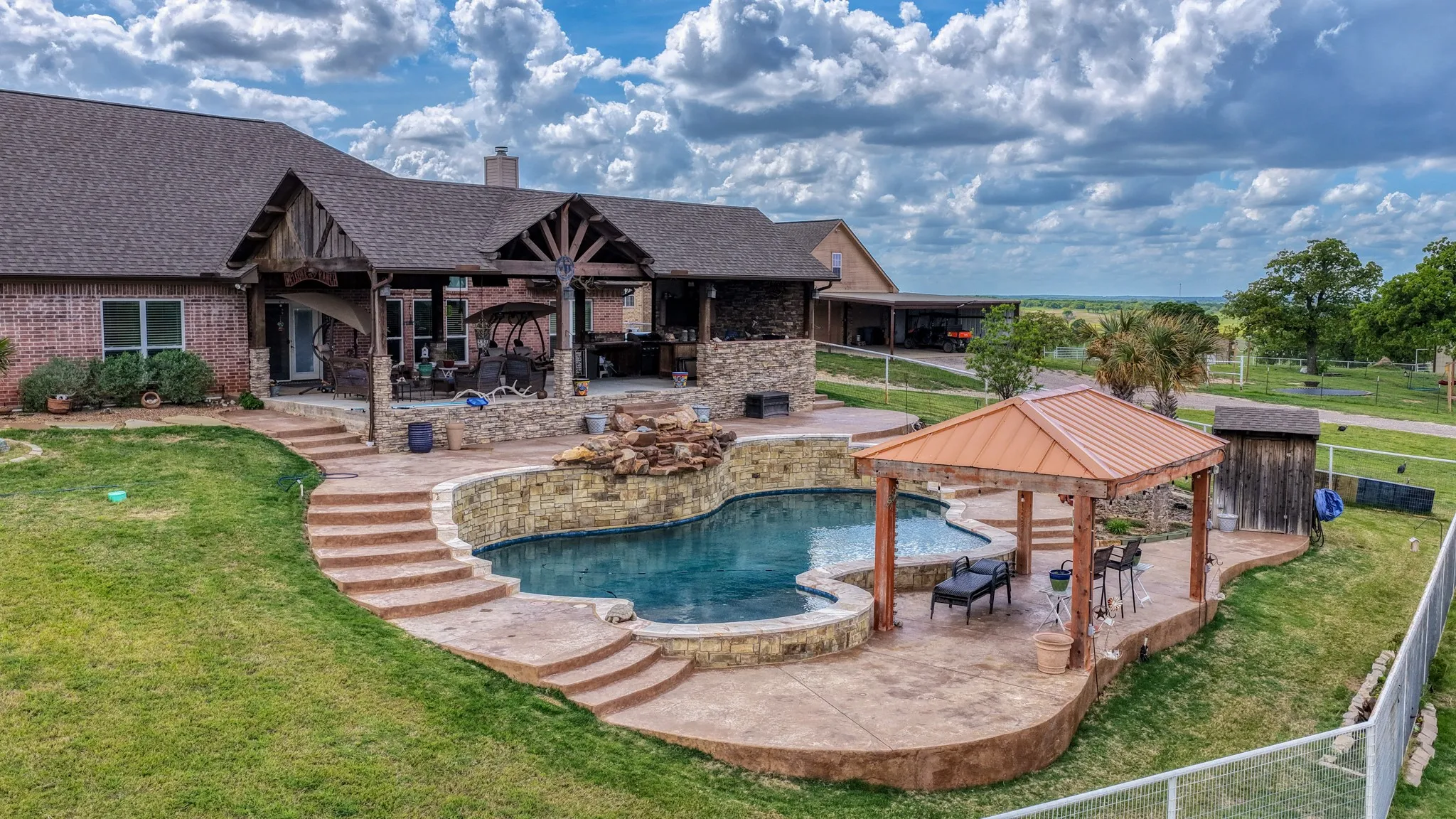 Expansive backyard featuring a freeform swimming pool with a rock waterfall feature, a covered patio with exposed beam accents, and a separate gazebo with a metal roof