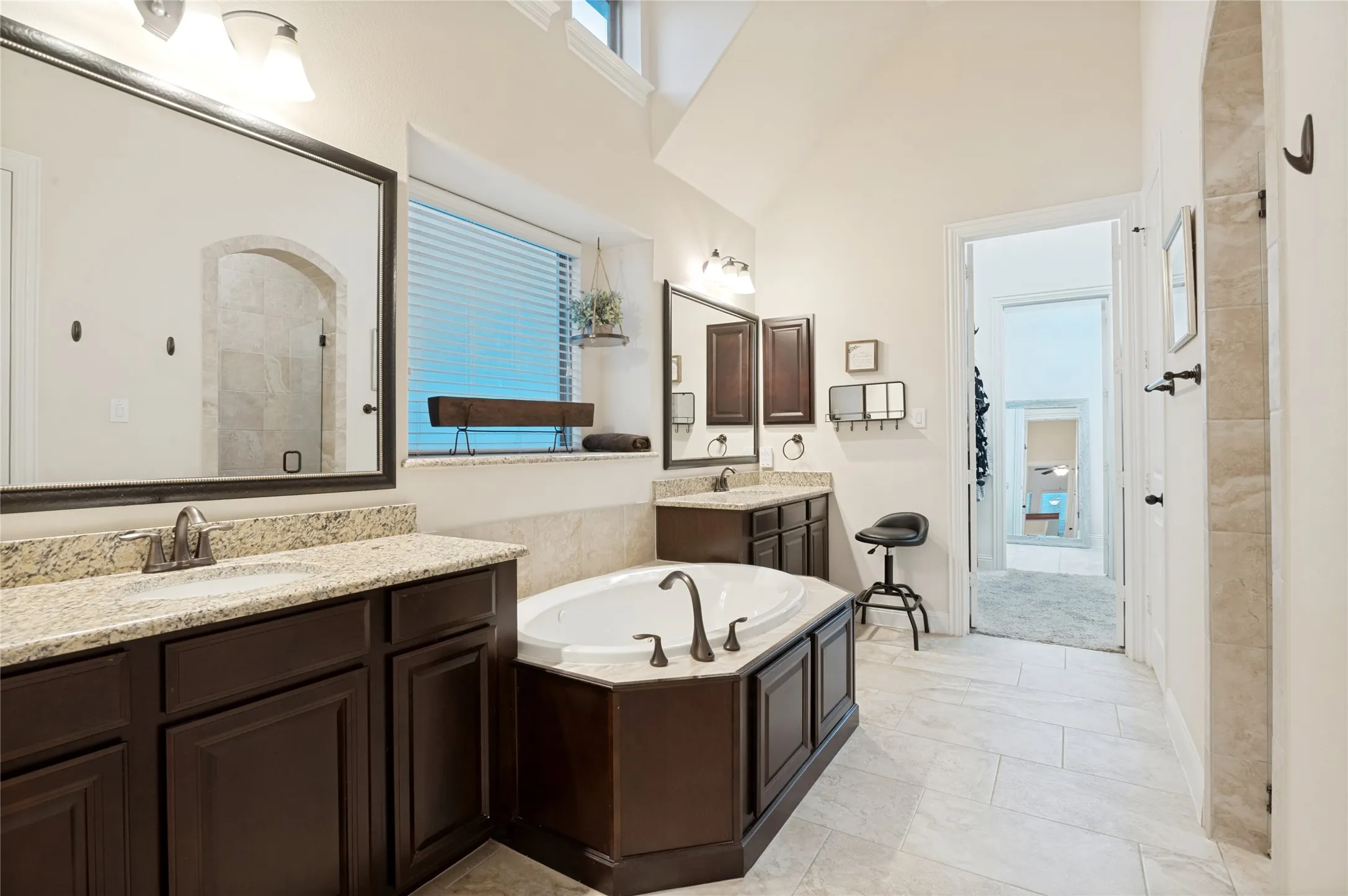 Spacious bathroom featuring a built-in soaking tub, dual vanity with granite countertops, dark wood cabinetry, and neutral tile flooring