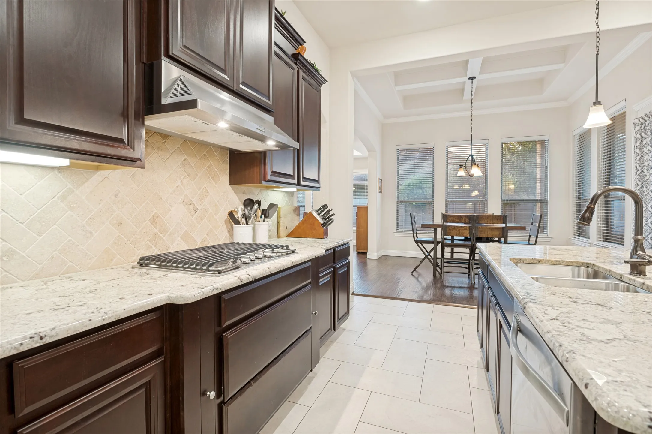 Gourmet kitchen featuring dark wood cabinetry, light-toned stone countertops, herringbone-patterned tile backsplash, a built-in gas range, and a stainless steel range hood