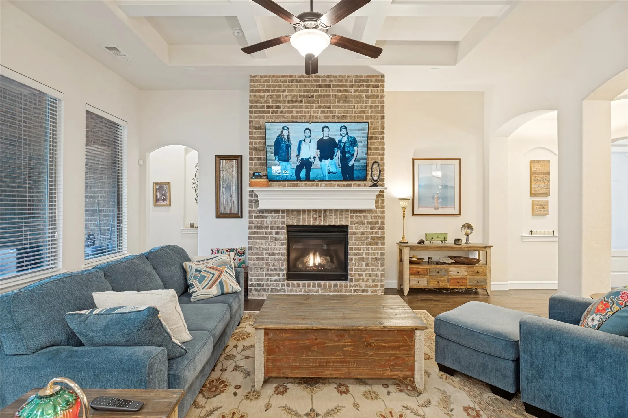 Living area featuring a brick-faced fireplace with a white mantel, a ceiling fan, tray ceiling details, and arched doorways