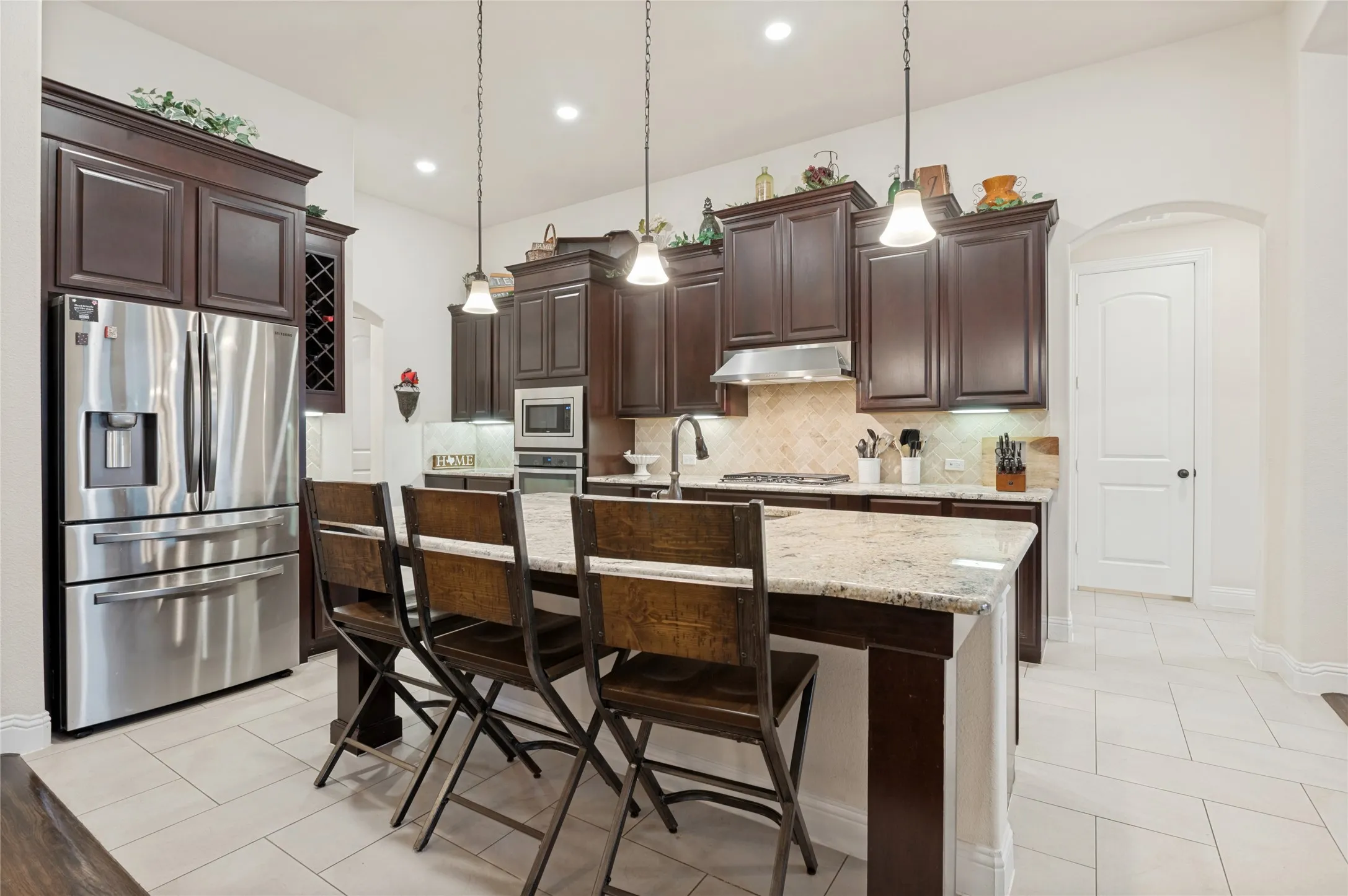 Kitchen featuring an island with stone countertops, dark wood cabinetry, stainless steel appliances, a tile backsplash, and light tile flooring