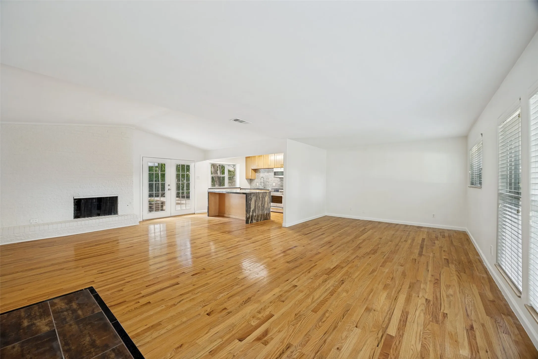 Spacious open-concept living area featuring wood-finish flooring, a white brick fireplace, vaulted ceilings, and double French doors