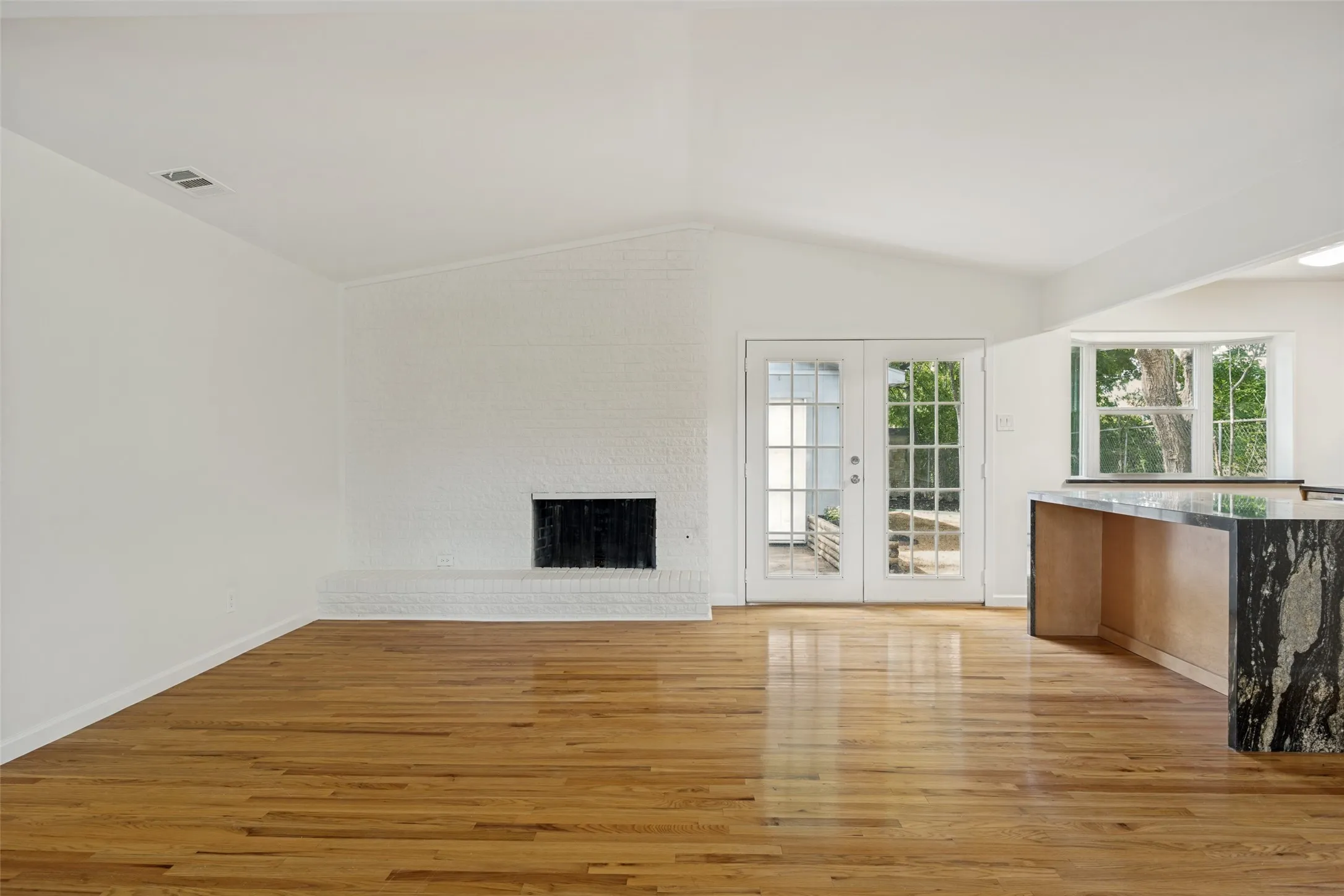Spacious interior featuring wood-finish flooring, a white brick fireplace, vaulted ceiling, and French doors