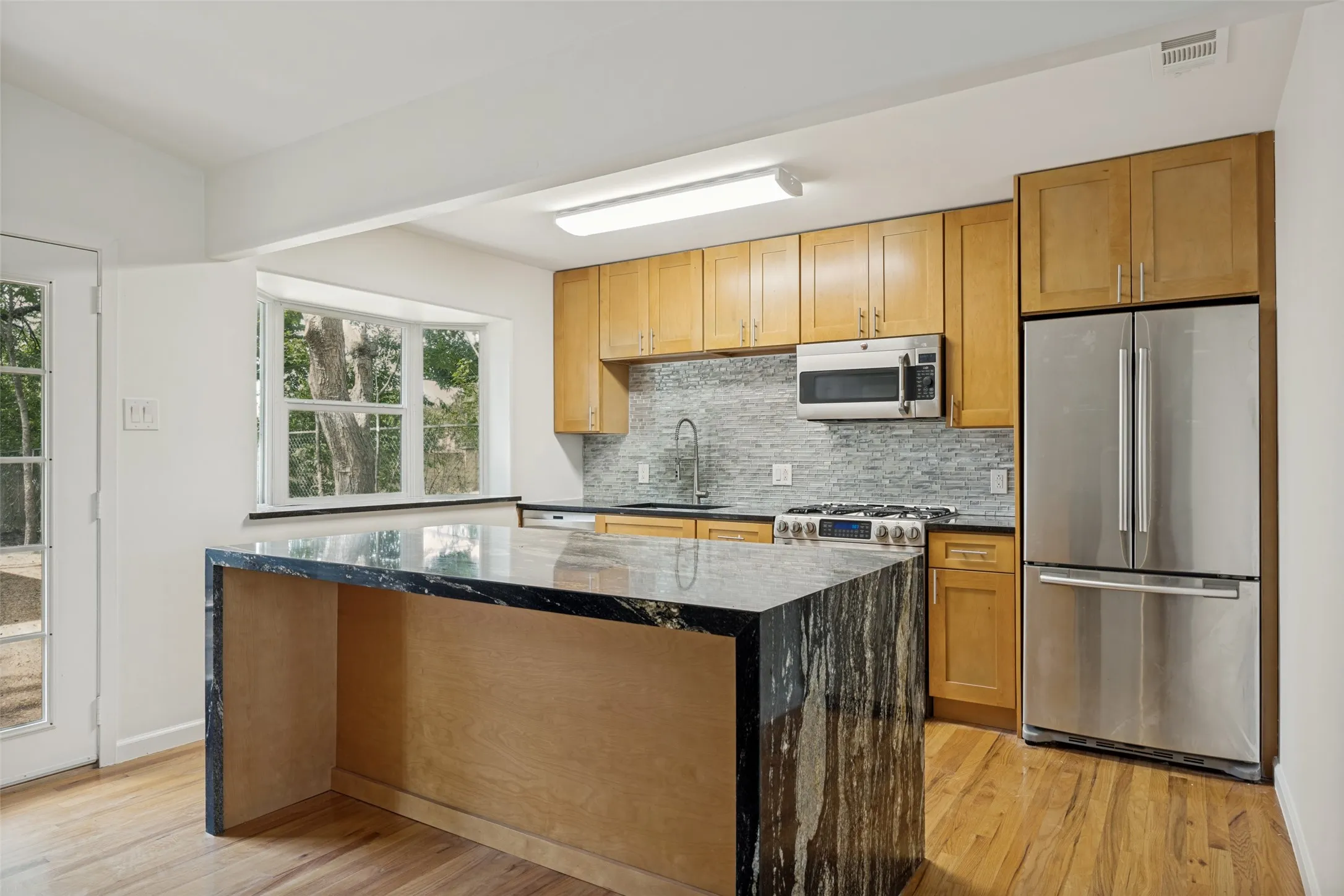 Kitchen featuring a large waterfall island with a granite countertop, stainless steel appliances, wood-finish flooring, light wood cabinetry, and a subway tile backsplash