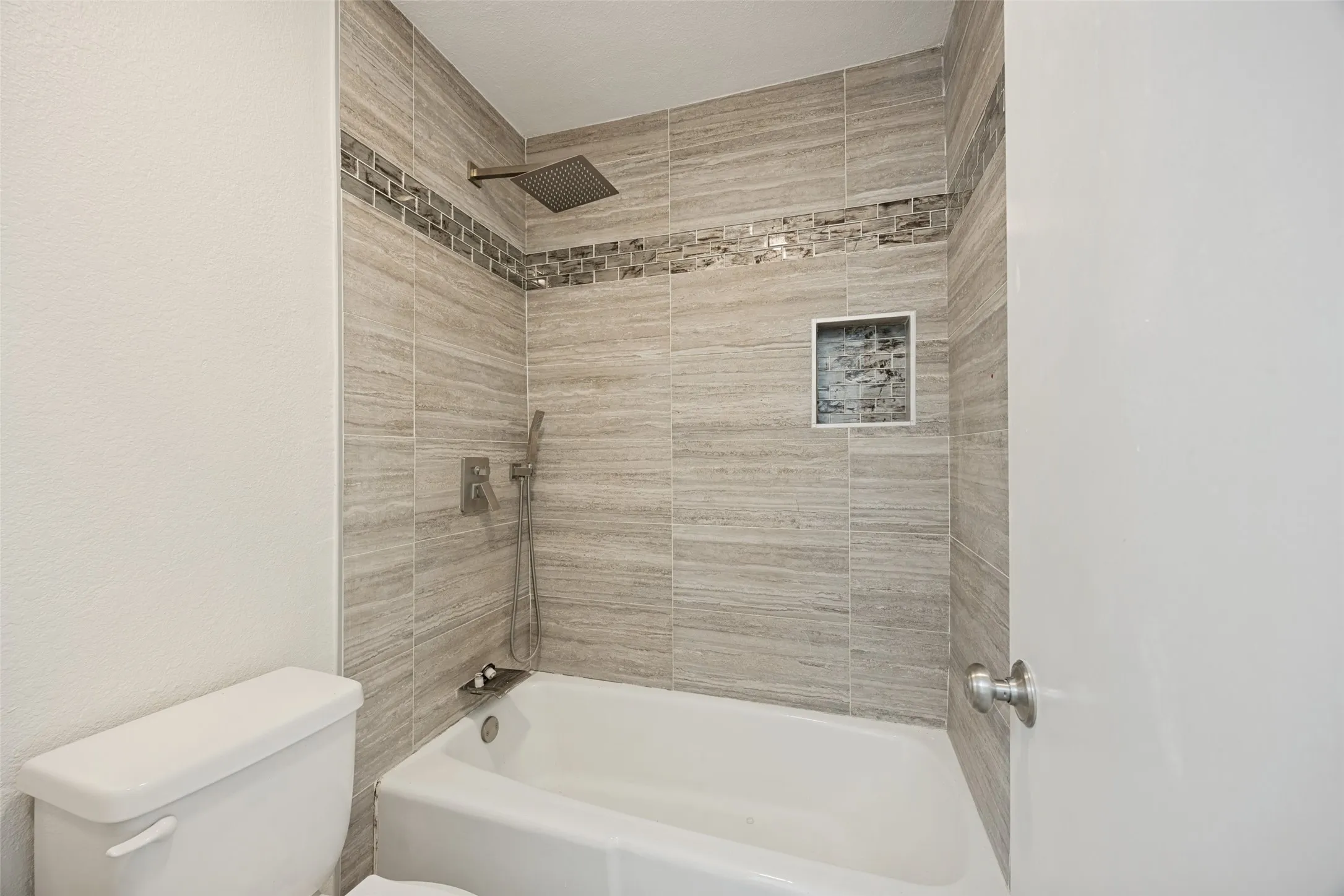 Bathroom featuring a tub/shower combination with large format tiled walls, a decorative mosaic tile accent band, a built-in shower niche, and a rain showerhead with a handheld sprayer