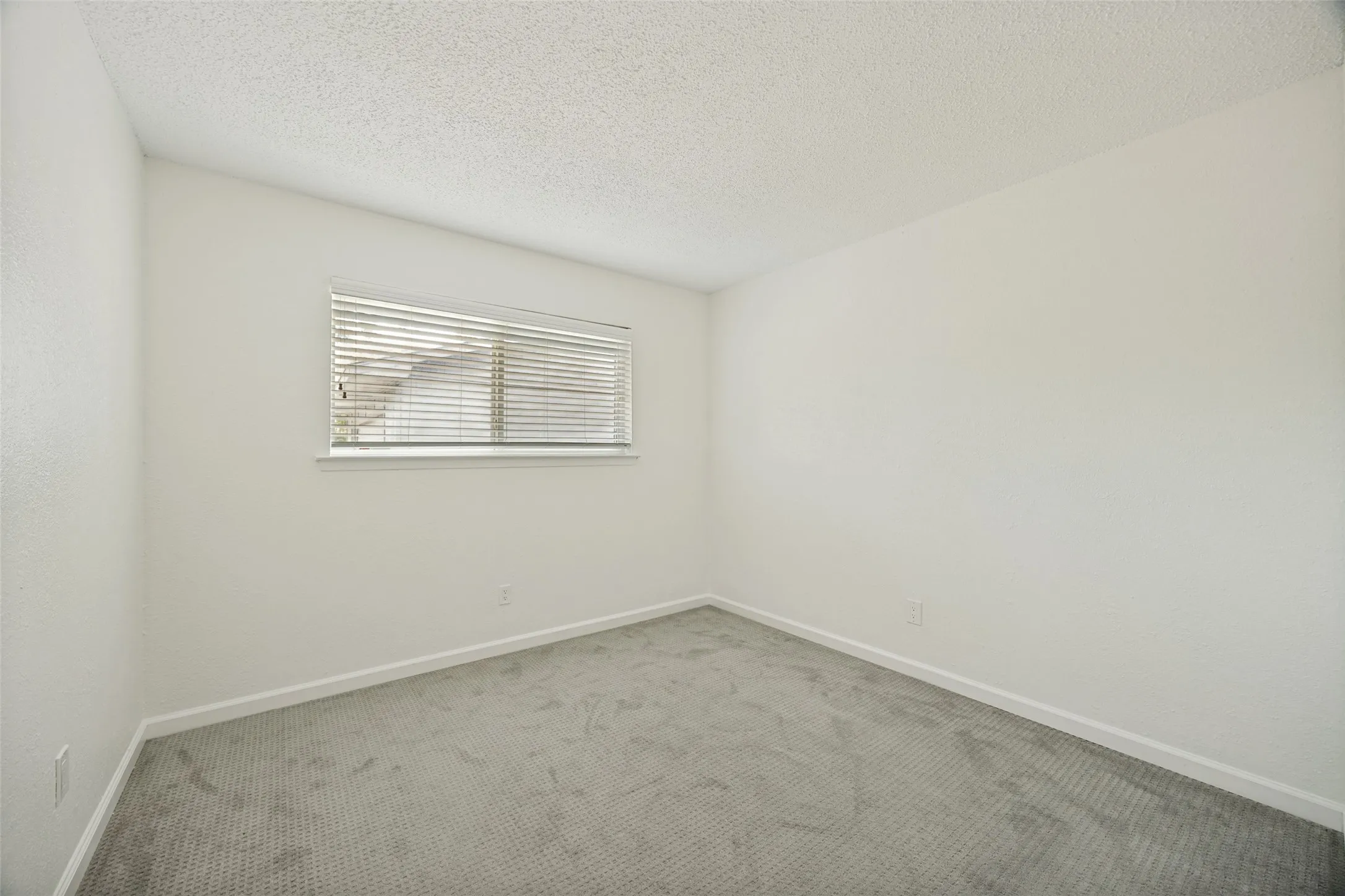 Neutral interior room featuring a window with horizontal blinds, light-toned walls, and a textured ceiling