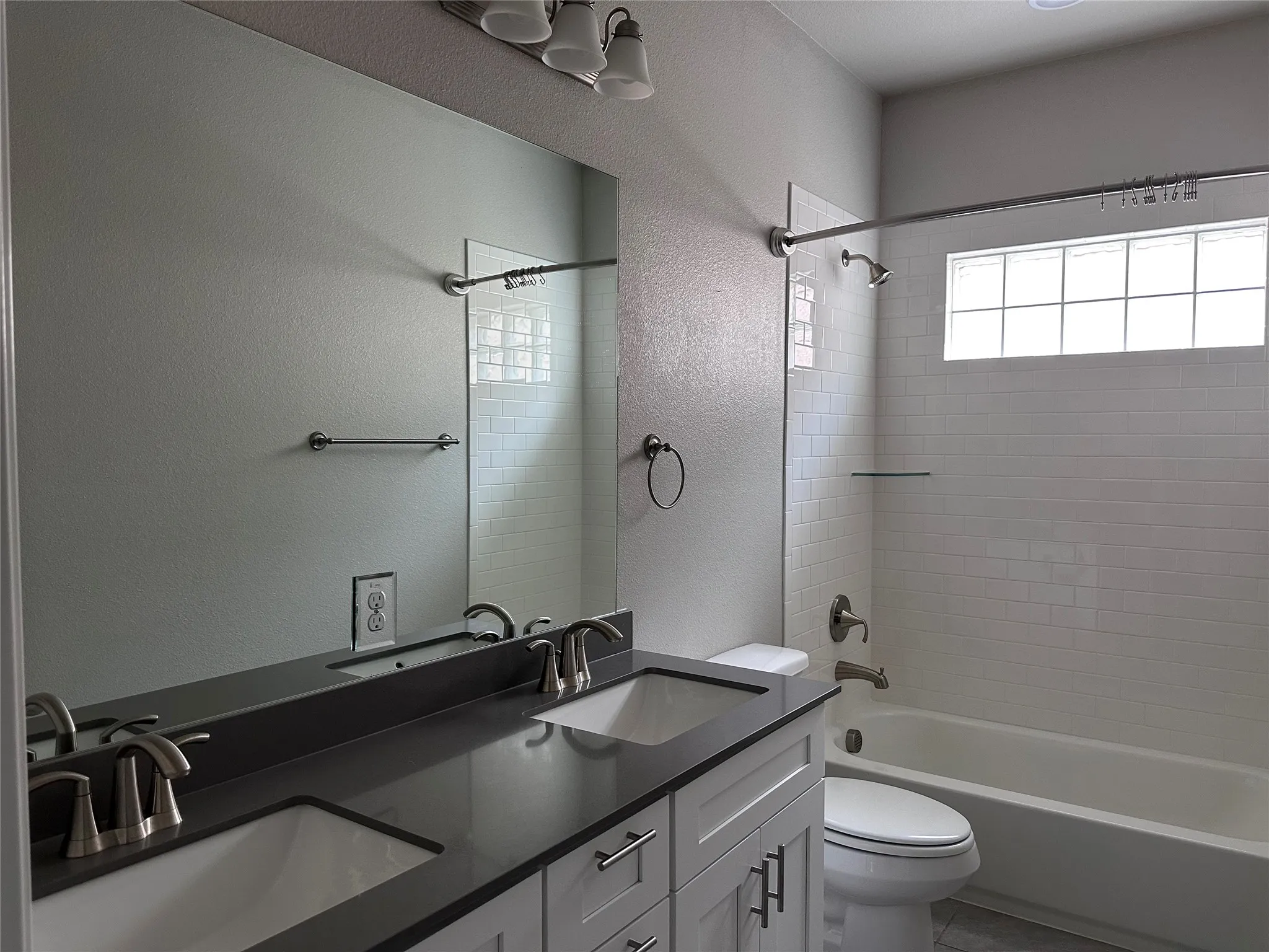Bathroom featuring a dual vanity with undermount sinks and dark countertops, white subway tile shower surround, glass block window, and brushed nickel fixtures