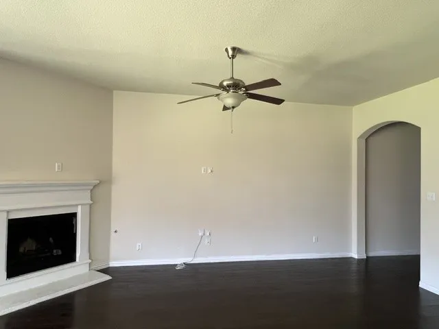 Spacious interior featuring dark wood-finish flooring, a white mantel fireplace, a ceiling fan with integrated lighting, and an arched doorway