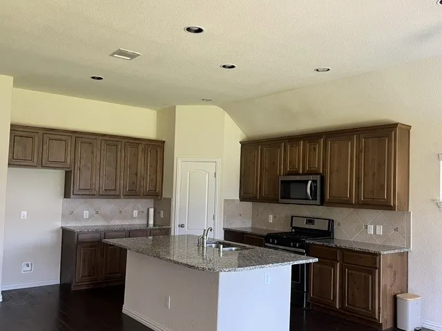 Kitchen featuring an island with a built-in sink, granite countertops, wood-finish cabinetry, stainless steel appliances, and dark wood-finish flooring