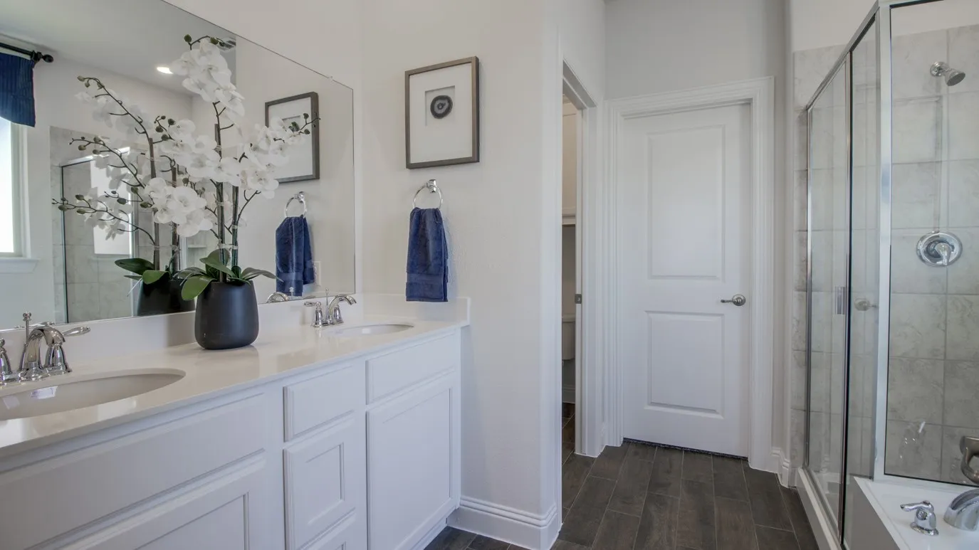 Bathroom featuring a dual vanity with white cabinetry and light-toned countertops, a full-width mirror, a glass-enclosed shower, and wood-finish flooring