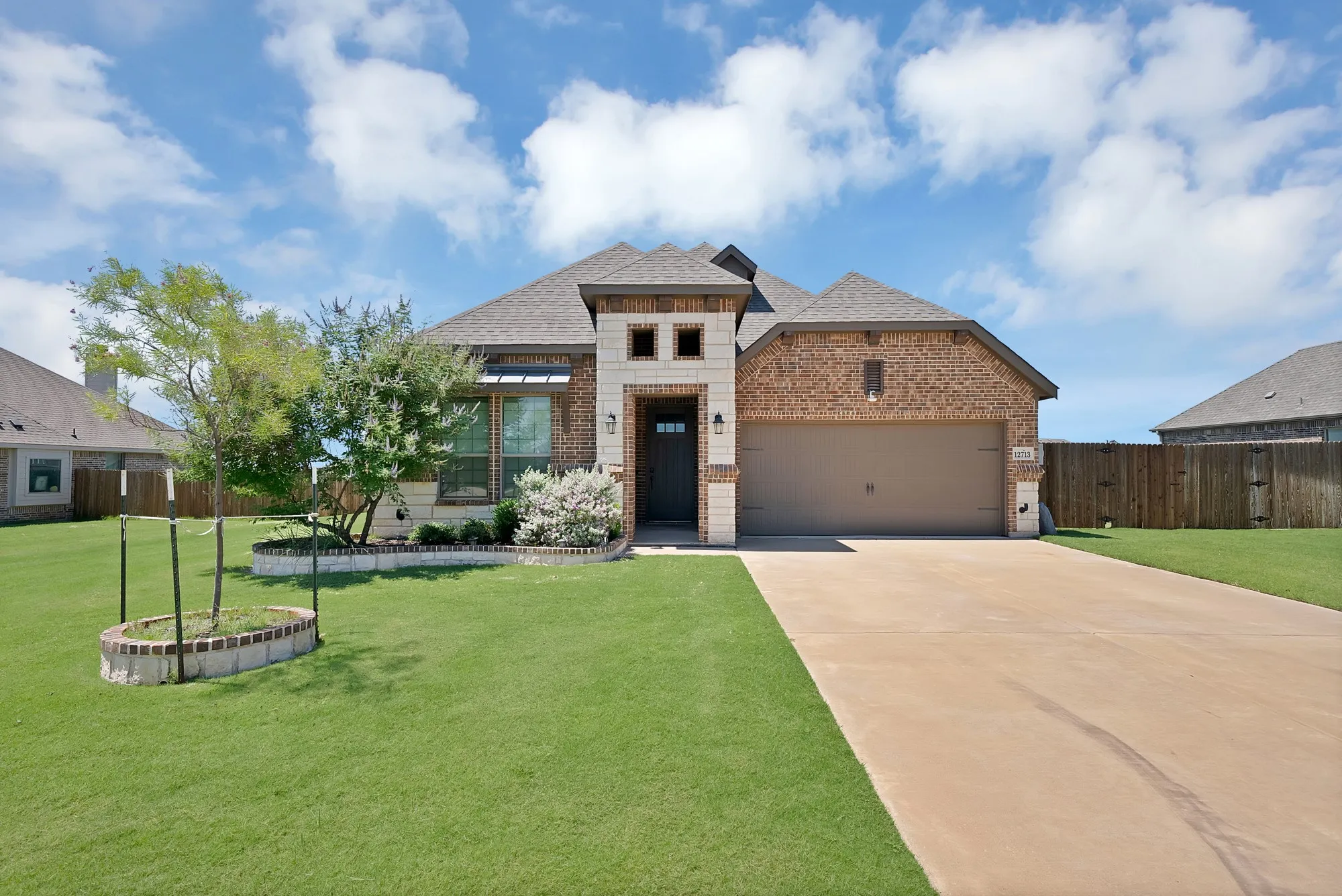 Brick and stone exterior featuring an attached two-car garage, a concrete driveway, and a front lawn with mature trees and landscaping