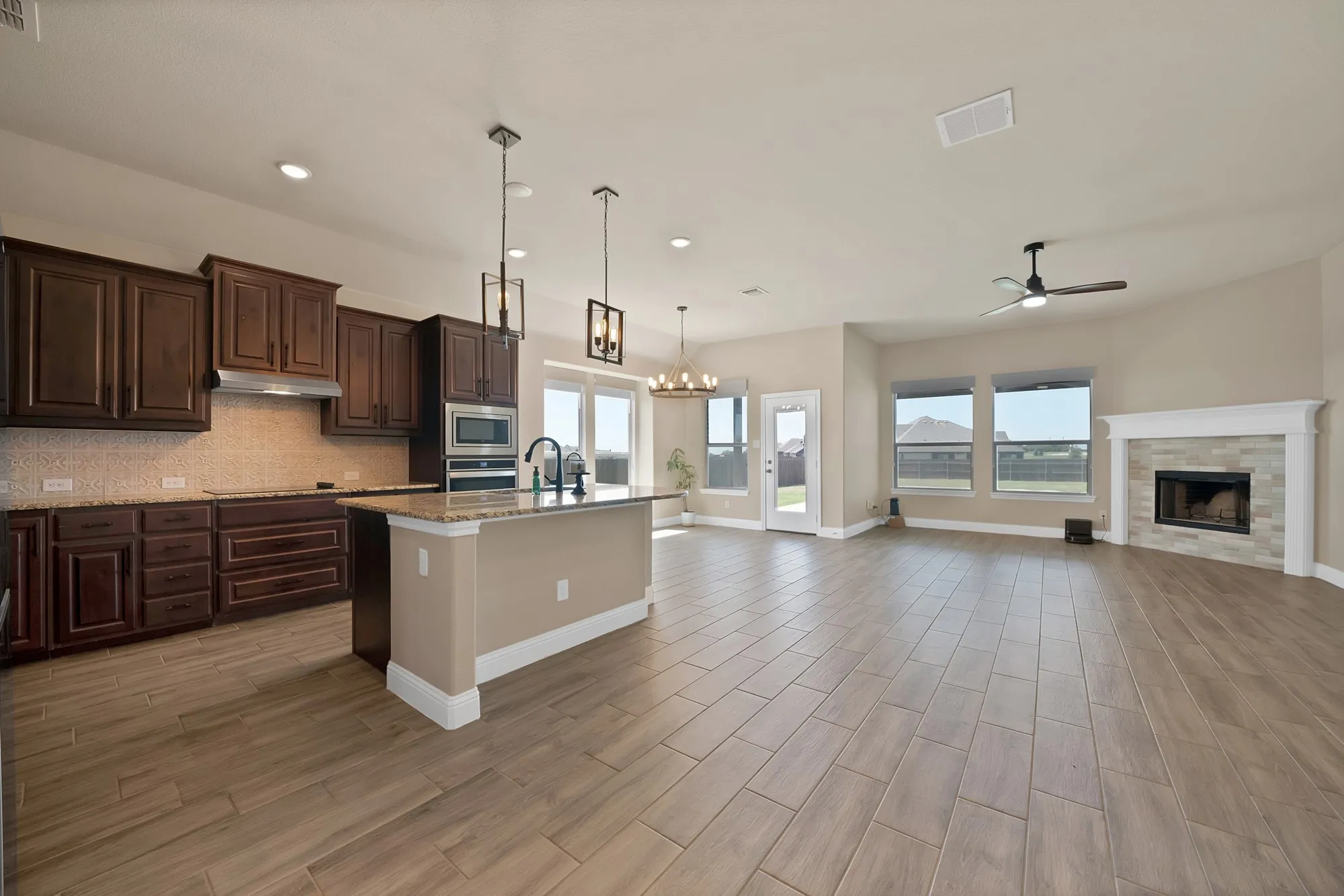 Open-concept living space featuring wood-finish tile flooring, a kitchen island with pendant lighting, dark wood cabinetry, and a fireplace with a stone surround