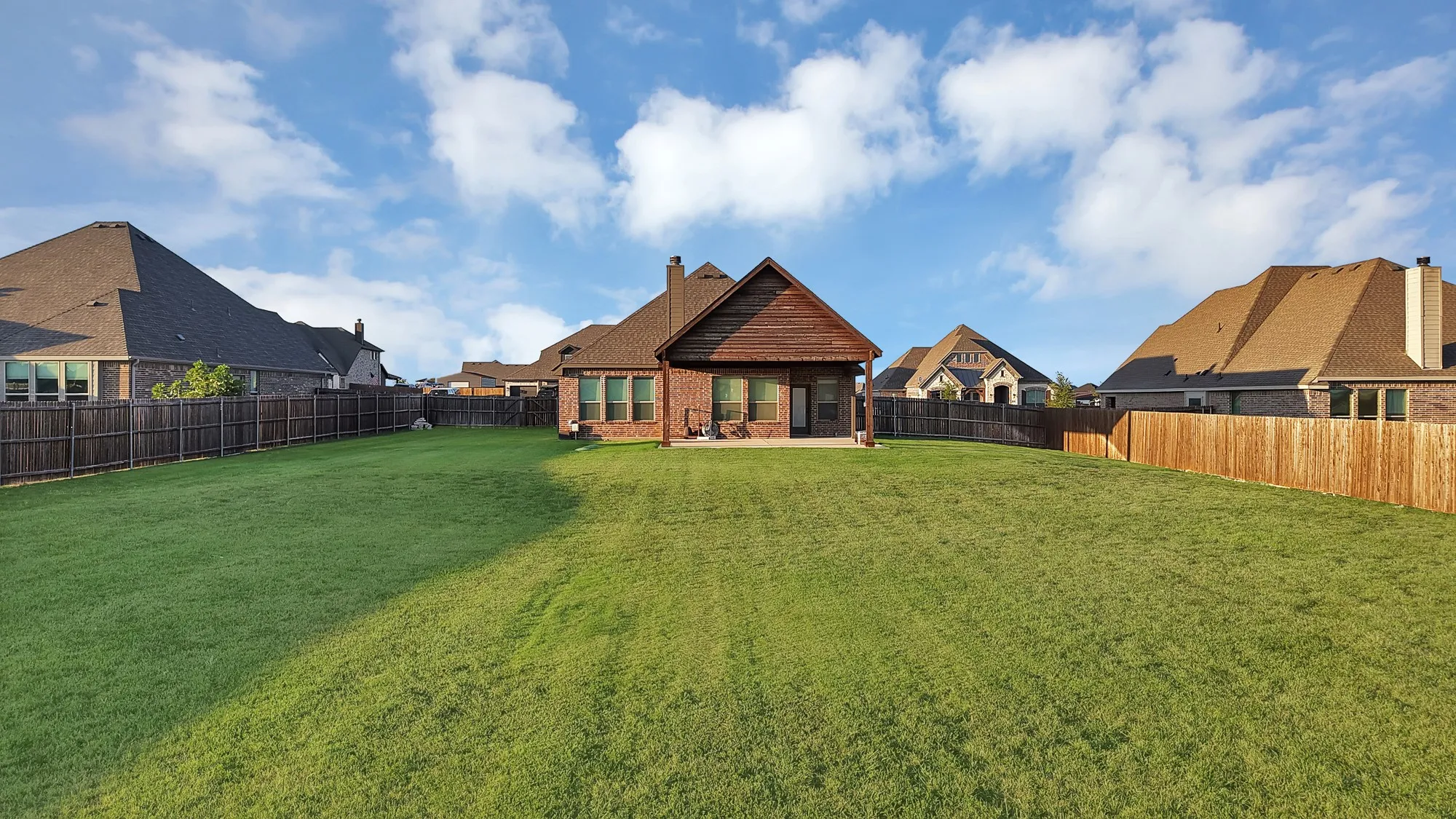 Expansive backyard featuring a large green lawn, full privacy fencing, a covered patio, and a brick exterior with wood siding accents