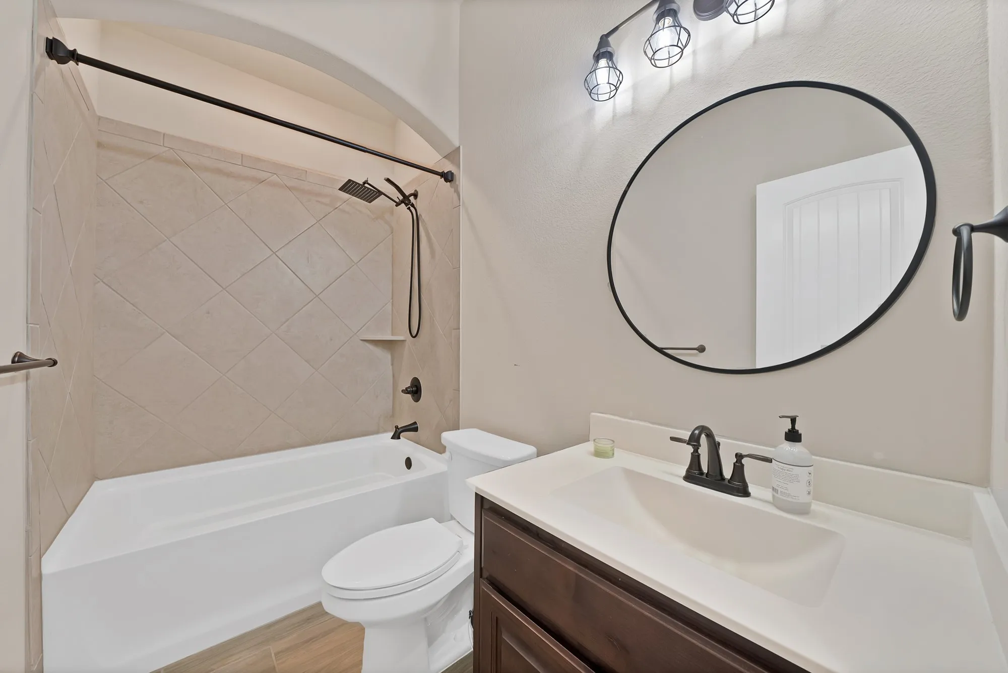 Bathroom featuring a combination shower and tub with diagonal tile surround, a dark wood-finish vanity with a single basin sink, a circular black-framed mirror, and a three-light fixture with caged accents