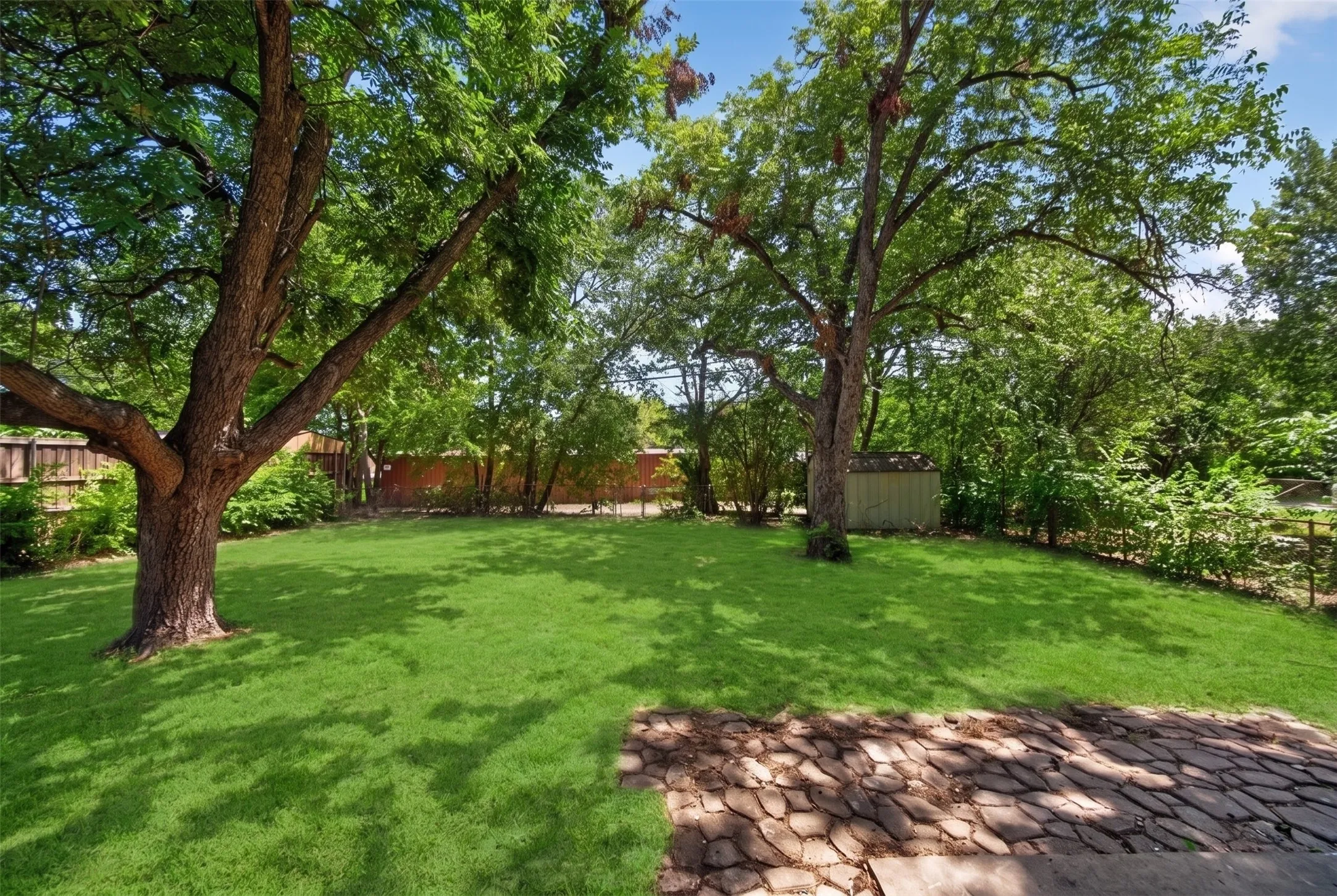 Expansive backyard featuring a manicured lawn, mature shade trees, stone patio, and a storage shed