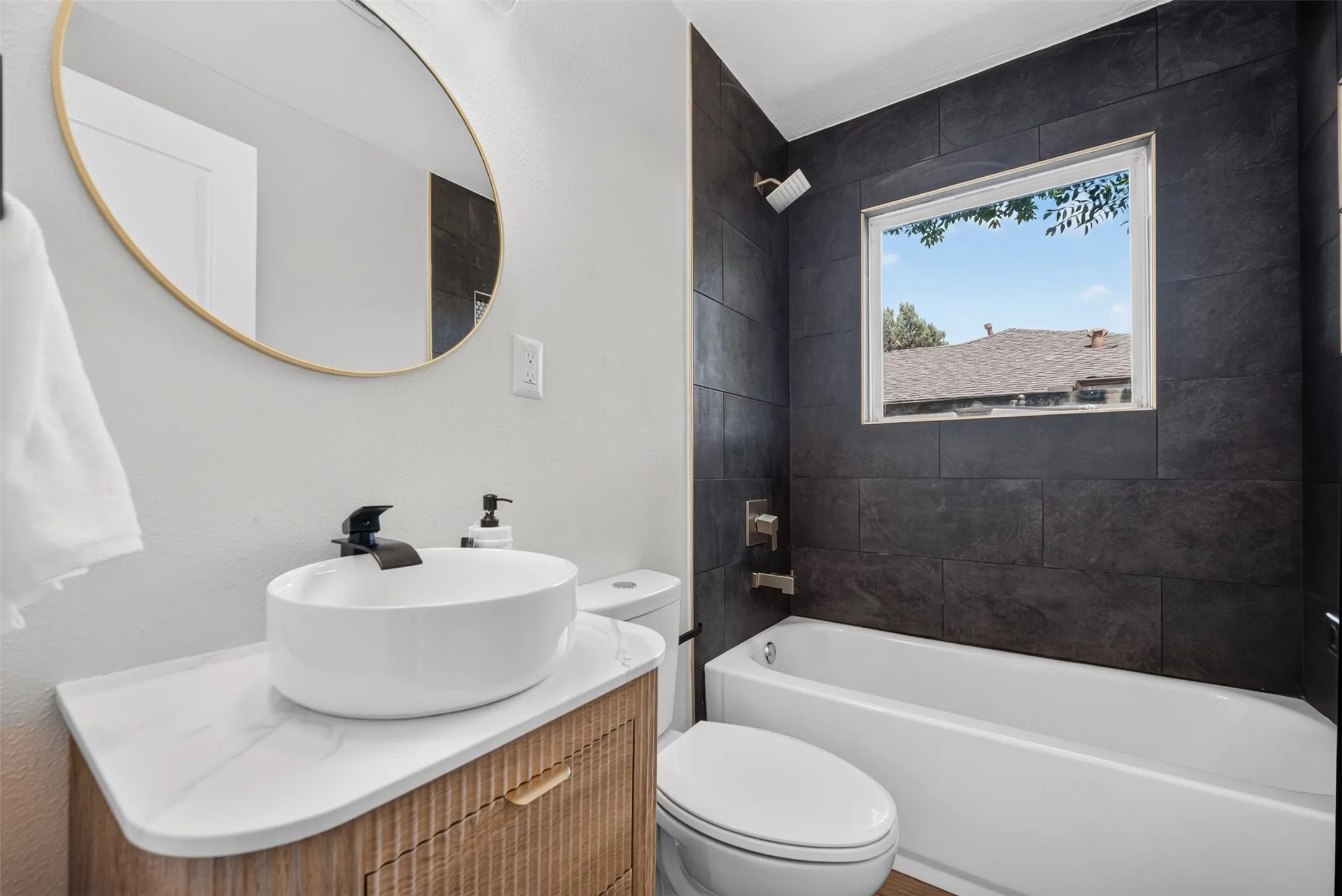 Bathroom featuring a modern vanity with a vessel sink, a gold-framed oval mirror, a white bathtub, and dark tile shower surround