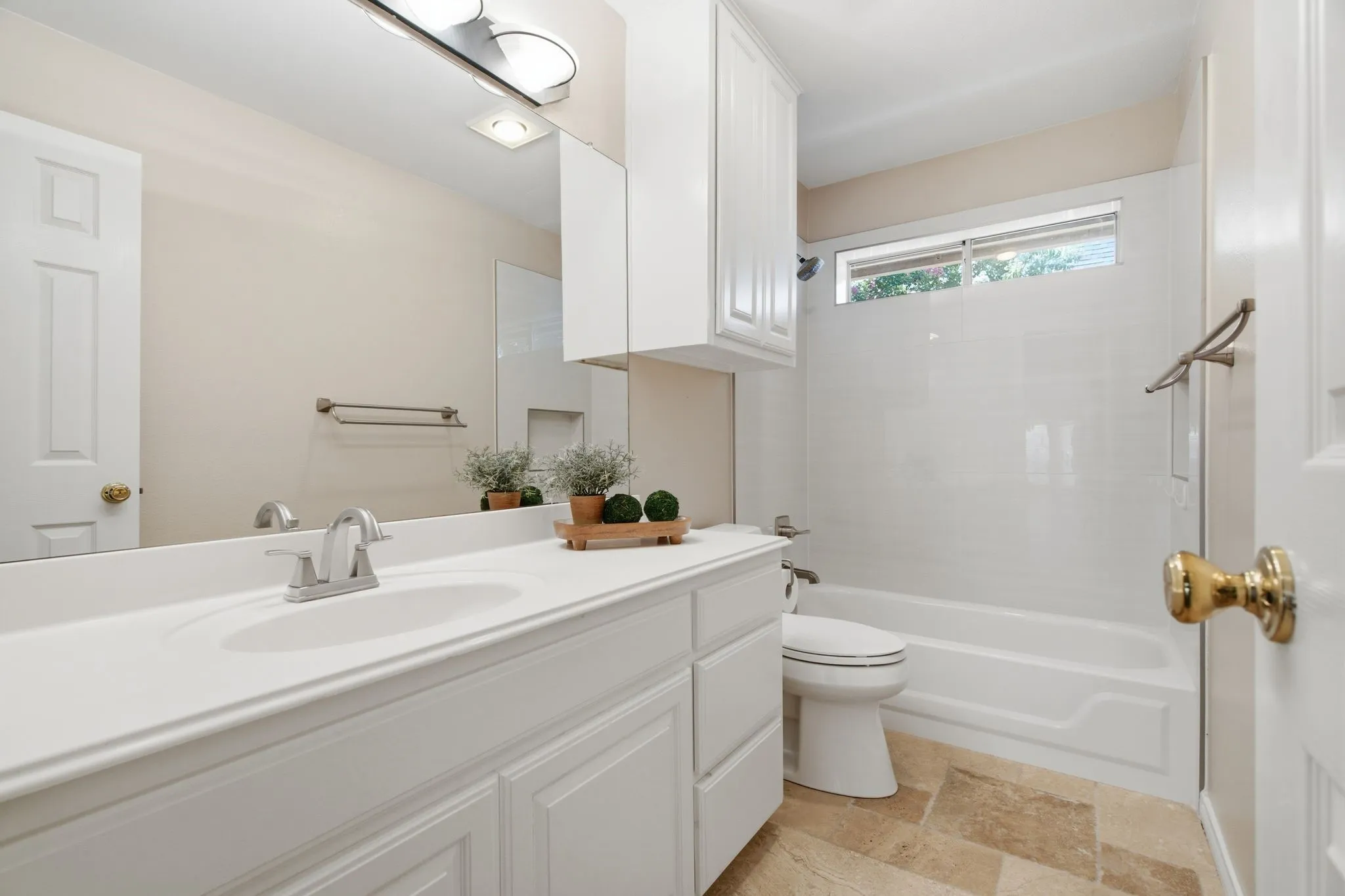 Bathroom featuring a single vanity with an integrated sink, white cabinetry, a bathtub with a shower, a horizontally-oriented window, and natural stone tile flooring