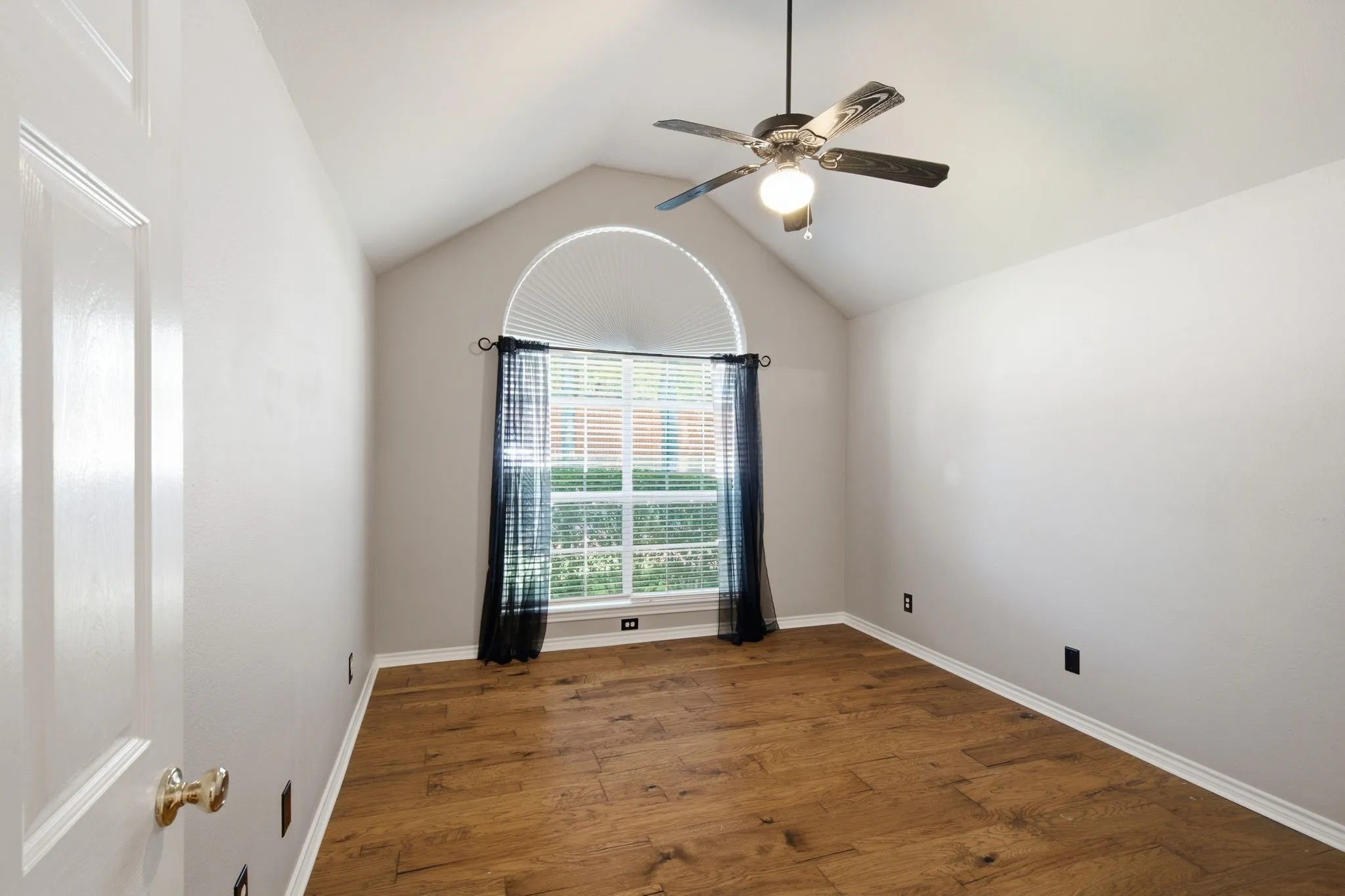 Vaulted ceiling room featuring wood-finish flooring, a large arched window, and a ceiling fan with integrated lighting