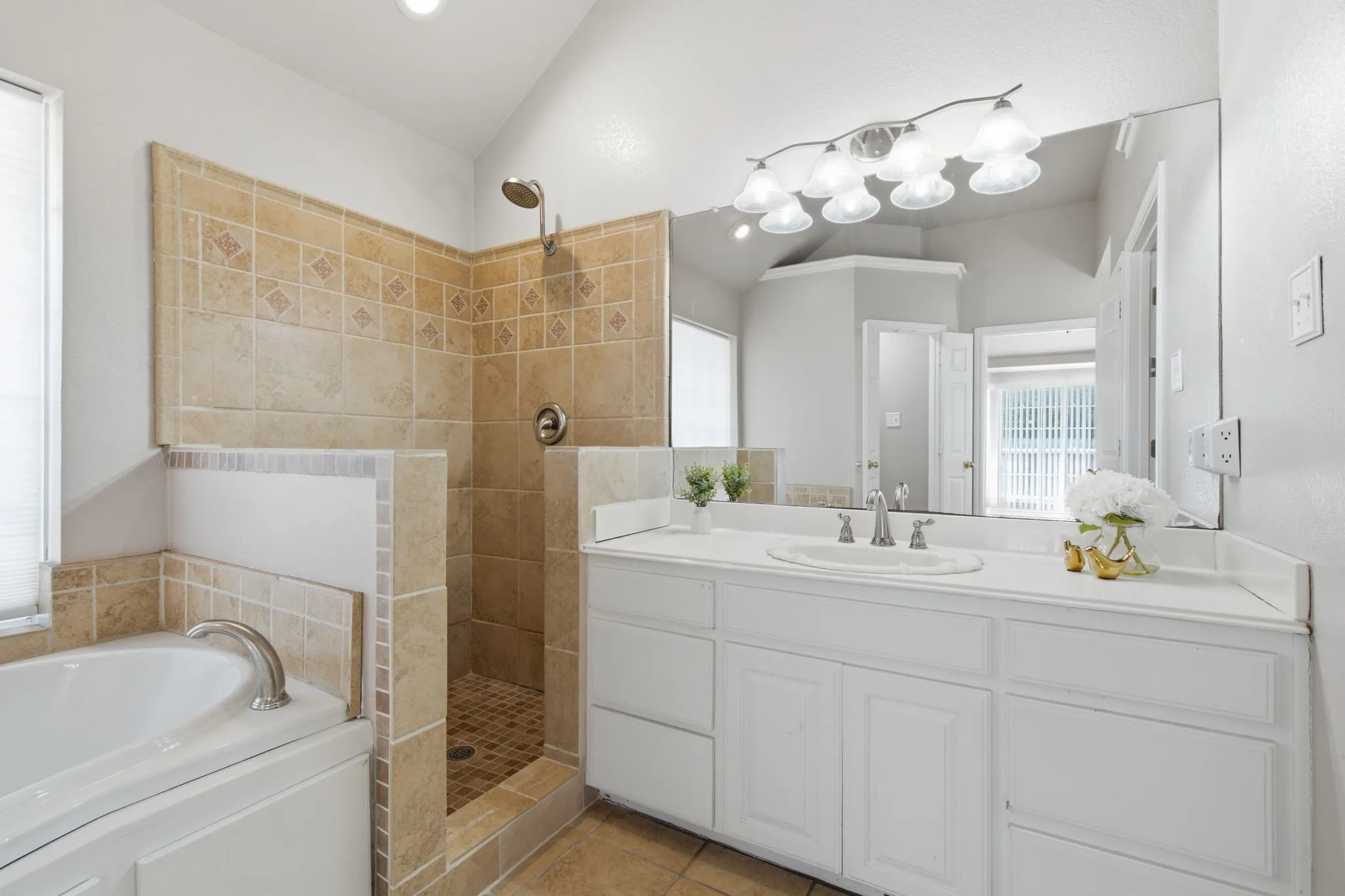 Bathroom featuring a neutral-toned tile shower with a built-in bench, a separate bathtub, a white vanity with an integrated sink, a large wall-mounted mirror, and a multi-bulb light fixture
