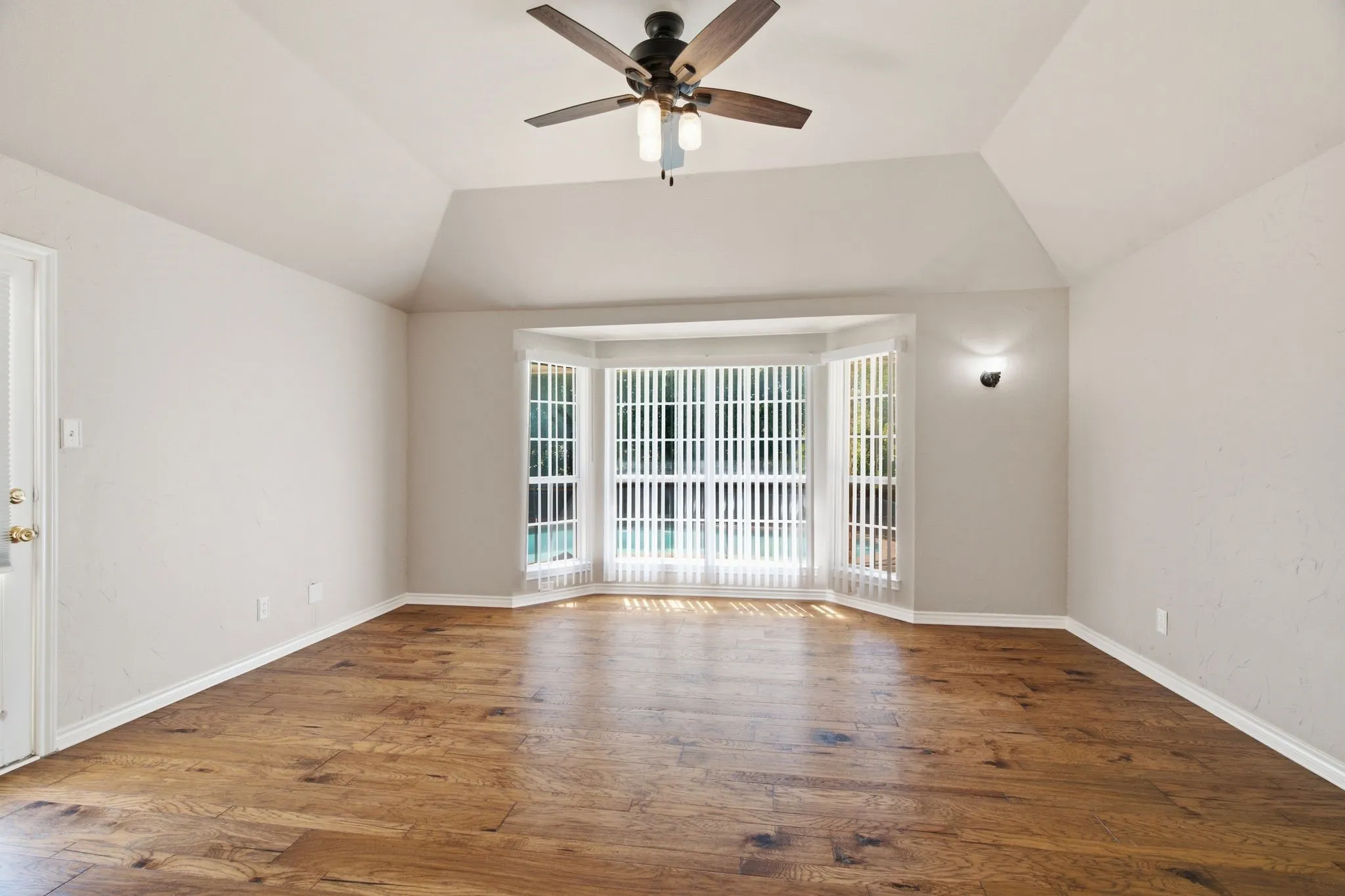 Spacious room featuring wood-finish flooring, a vaulted ceiling, a ceiling fan with integrated lighting, and a prominent bay window with vertical blinds