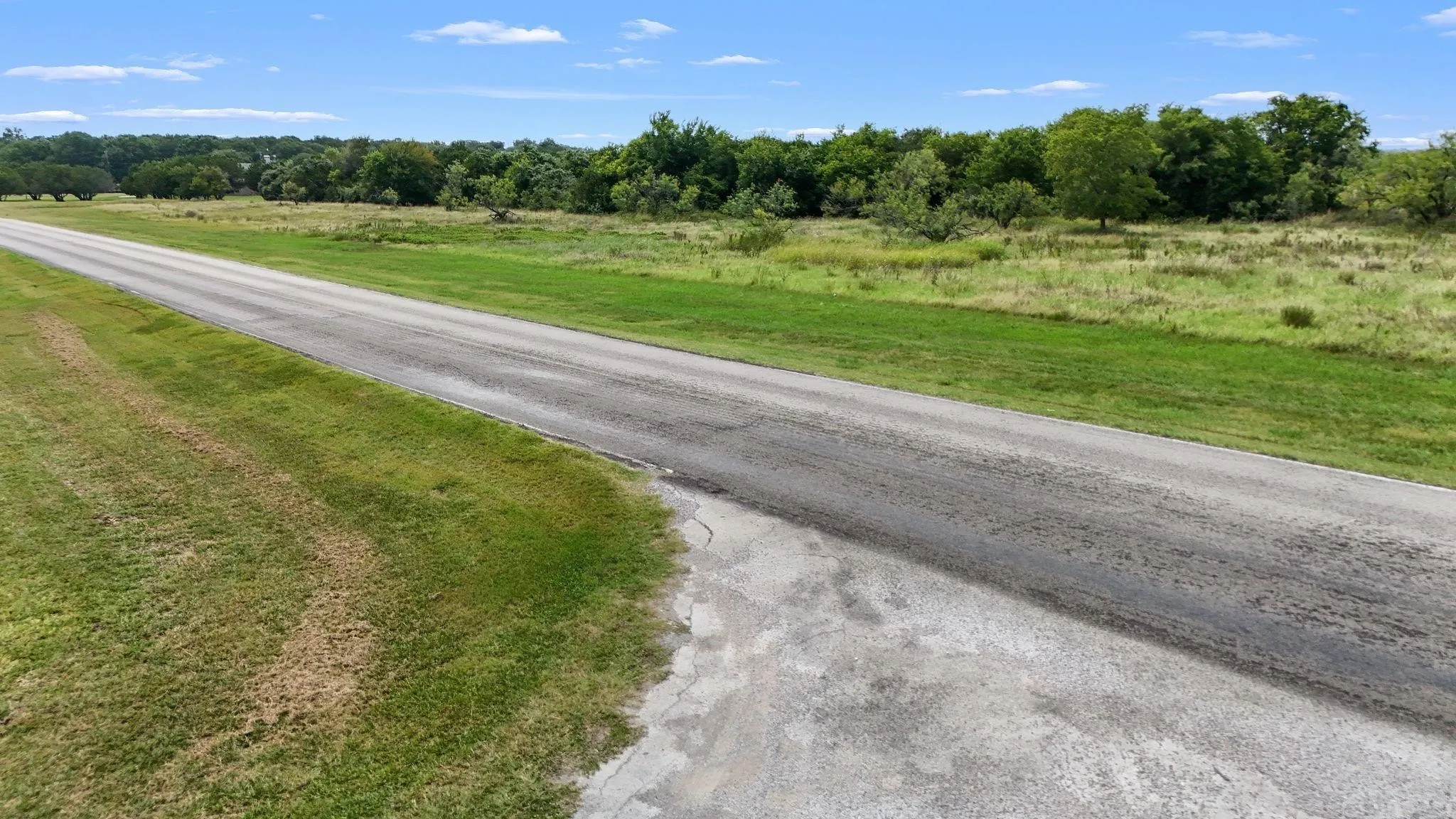 Expansive land parcel featuring a long paved road, mature tree line, and open grassy areas under a clear sky