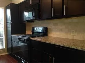 Kitchen featuring black cabinetry with brushed nickel hardware, a built-in microwave, gas range, dishwasher, and stone-finish countertops with a tile backsplash