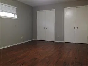 Spacious room featuring rich wood-finish flooring, neutral gray walls, two sets of white bifold closet doors, and white baseboards