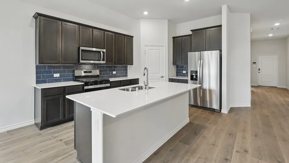 Modern kitchen featuring an expansive island with a double basin sink, dark wood cabinetry, stainless steel appliances, a blue subway tile backsplash, and light wood-finish flooring