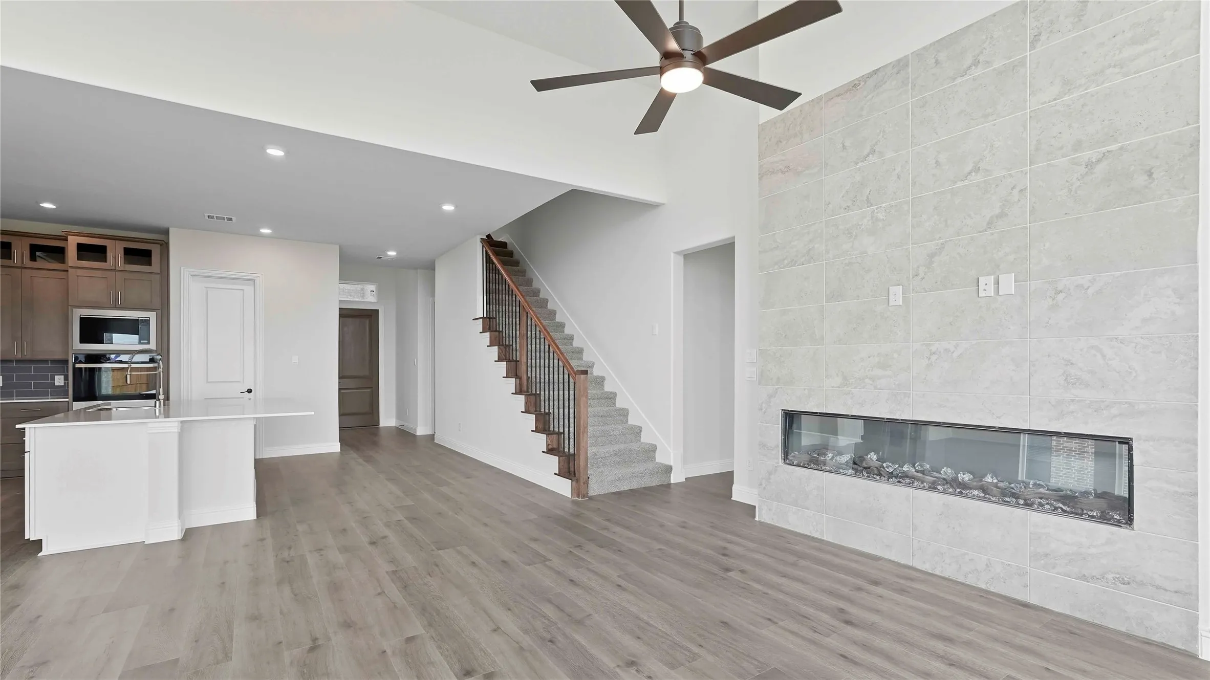 Open-concept living space featuring wood-finish flooring, a contemporary linear fireplace with a textured tile surround, and a ceiling fan