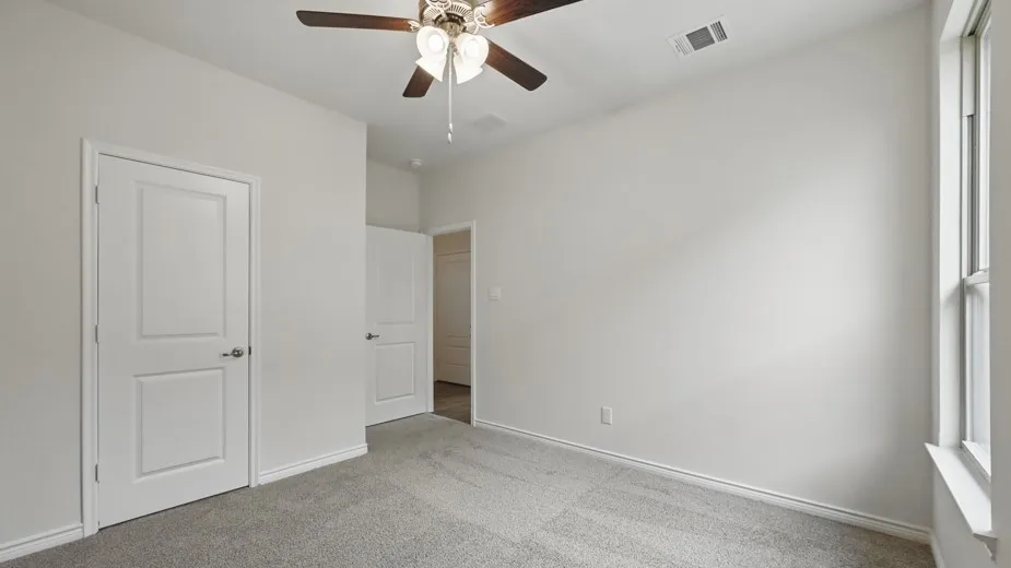 Carpeted interior room featuring light gray walls, white trim, a ceiling fan with light fixture, and a window offering natural illumination