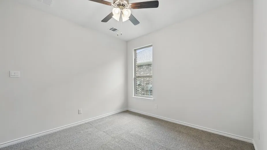 Carpeted interior room featuring a ceiling fan with light fixture, a window with exterior brick siding visible, and white baseboards