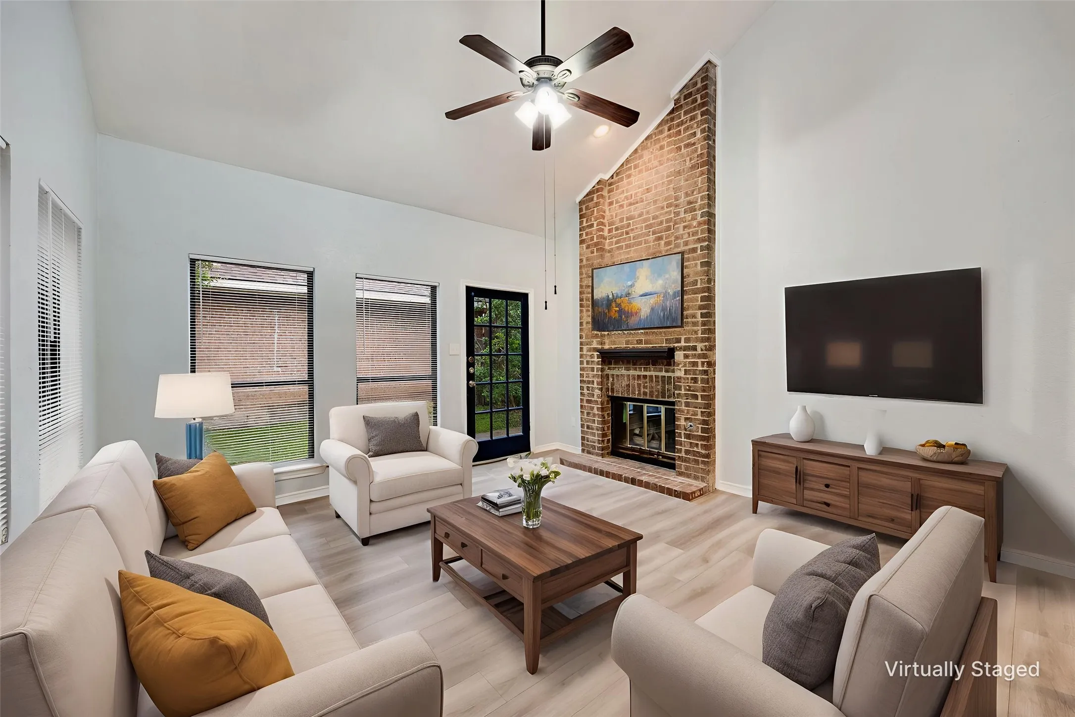 Living area with a high vaulted ceiling, light wood-finish flooring, a brick-faced fireplace, a ceiling fan with integrated lighting, and multiple windows