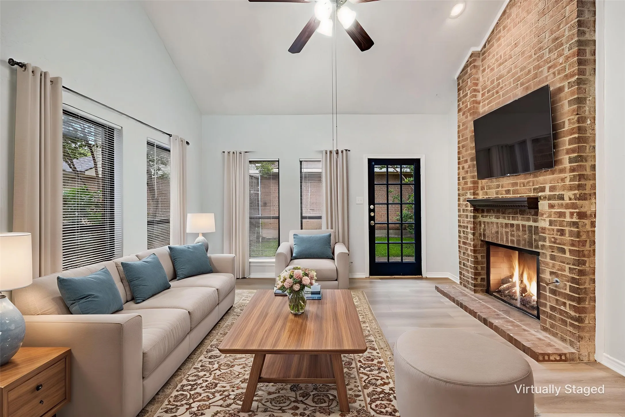 Living room featuring a brick fireplace with a wood mantel, vaulted ceiling, wood-finish flooring, and multiple windows with blinds