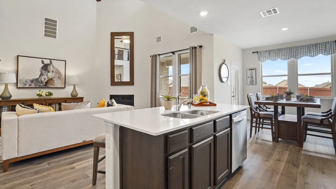 Open concept living area featuring a kitchen island with quartz countertop and stainless steel double sink, wood-finish flooring, a dining area with a window overlooking the exterior, and a living space with a vaulted ceiling and fireplace