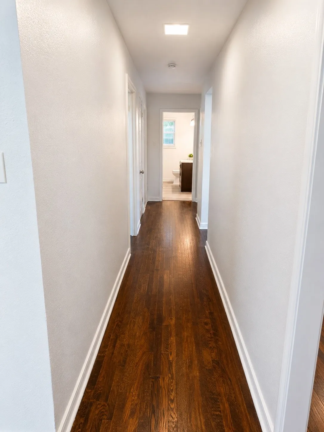 Hallway featuring rich wood-finish flooring, white baseboards, and bright overhead lighting