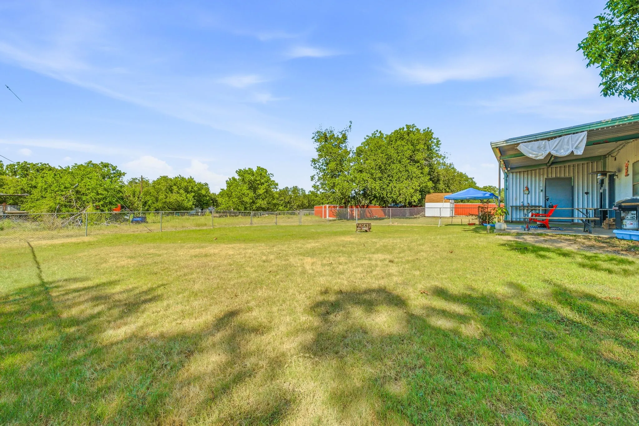 Expansive grassy yard with chain-link fencing, mature trees, and a covered patio featuring a metal roof
