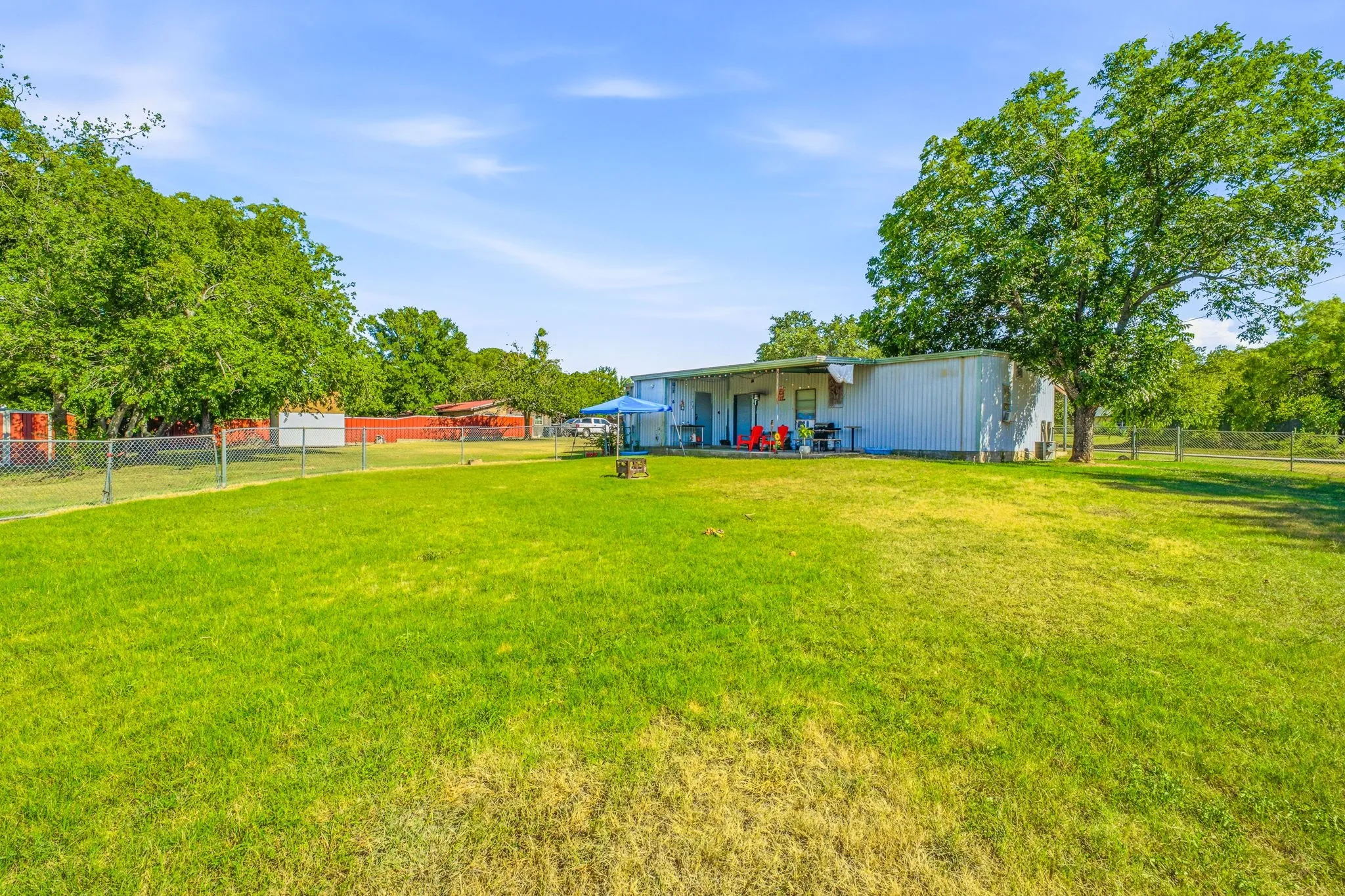 Expansive green lawn with a chain-link fence, mature trees, and a metal-sided structure featuring an extended covered patio