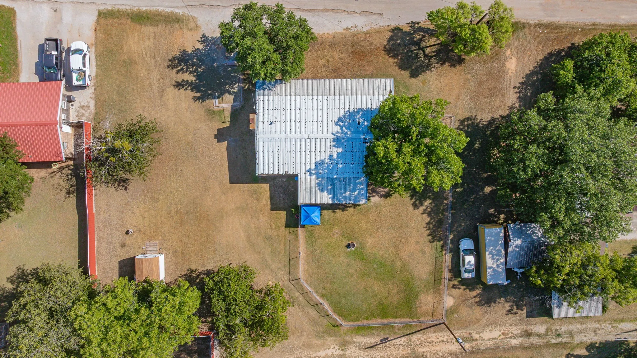 Aerial property overview featuring a main building with a metal roof, expansive unpaved lot, scattered mature trees, and an additional red-roofed structure