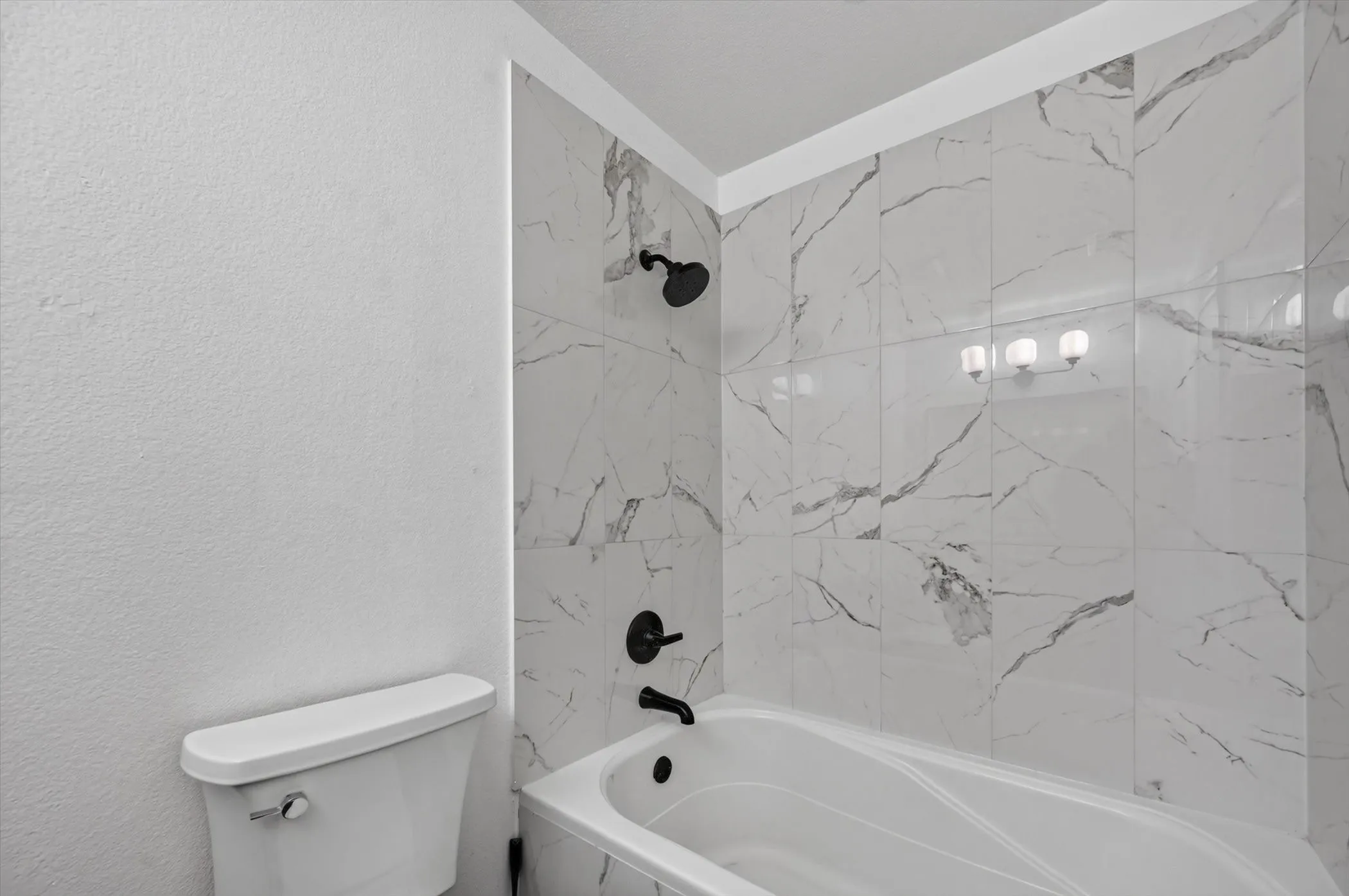 Bathtub with marble-look tile surround, matte black shower fixtures, and a three-light vanity fixture