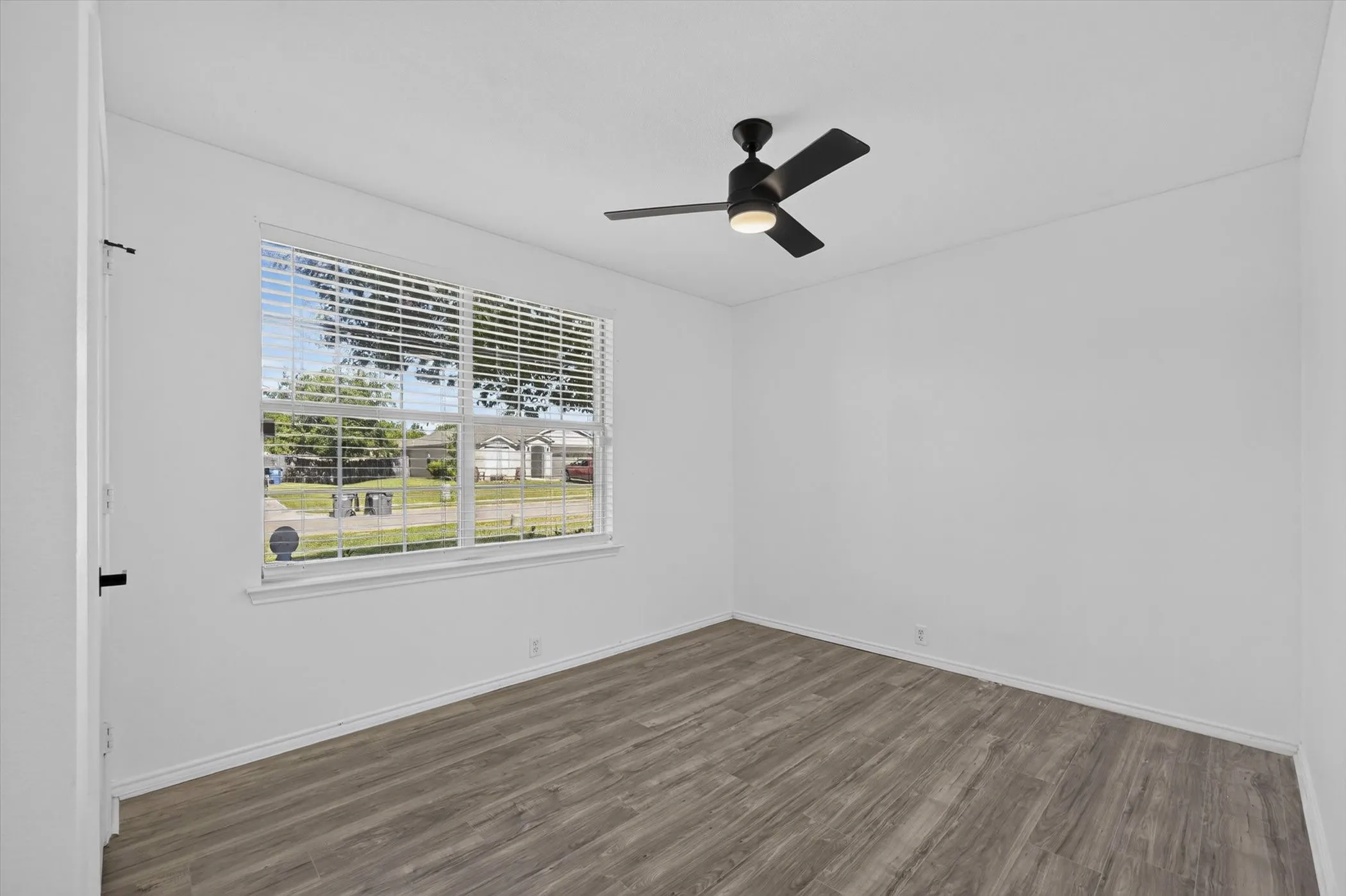 Spacious room featuring wood-finish flooring, a large window with blinds, a modern ceiling fan, and white baseboards